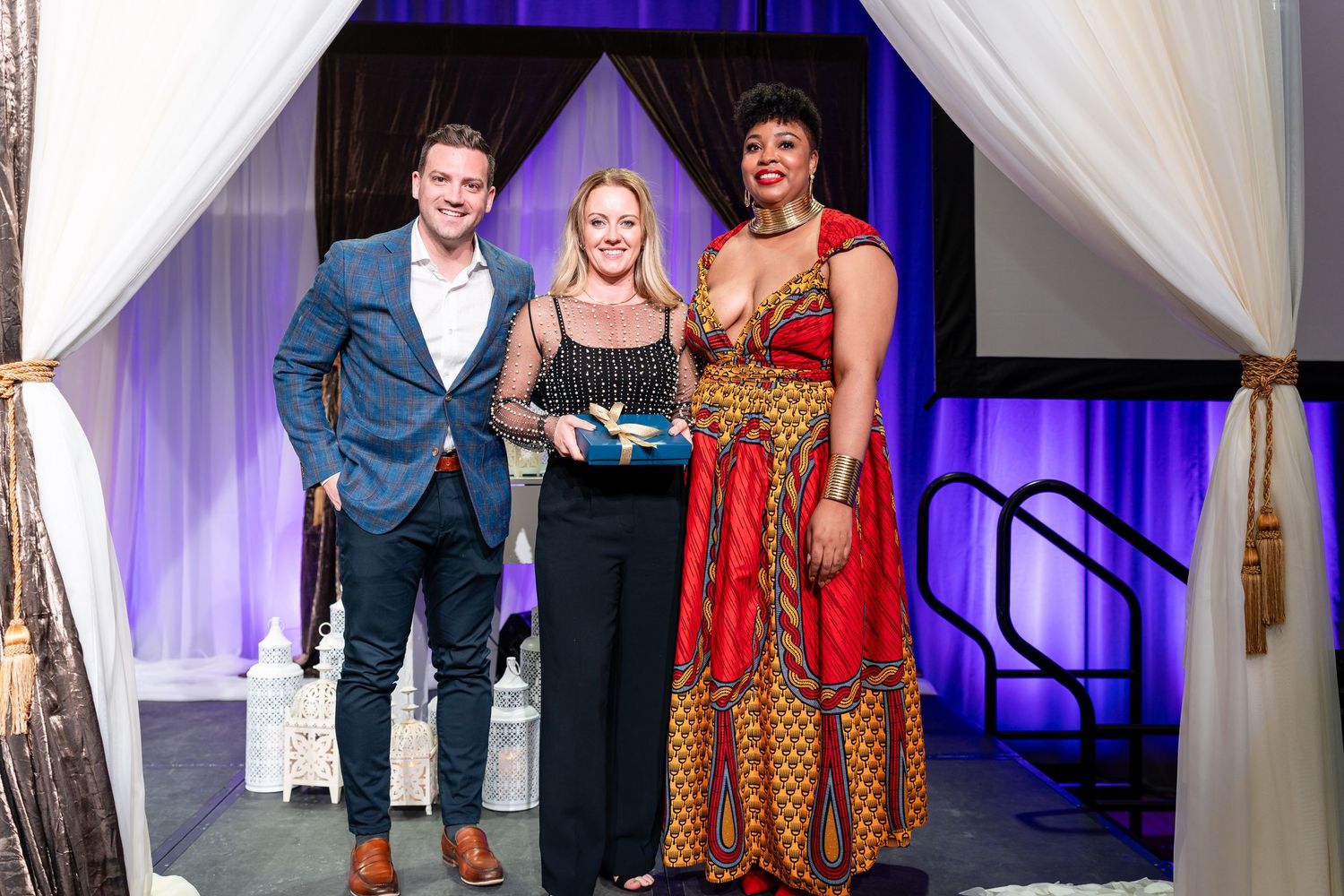 Three people stand under white drapes at an awards ceremony, with one wearing a vibrant patterned dress.