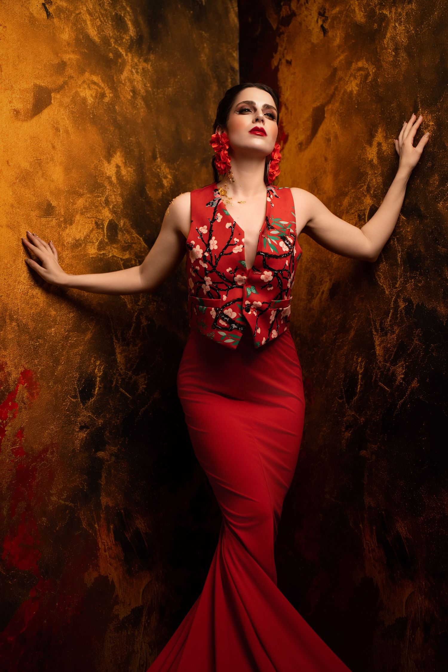 Dramatic fashion photo of woman in red mermaid dress with floral detail against textured golden wall.
