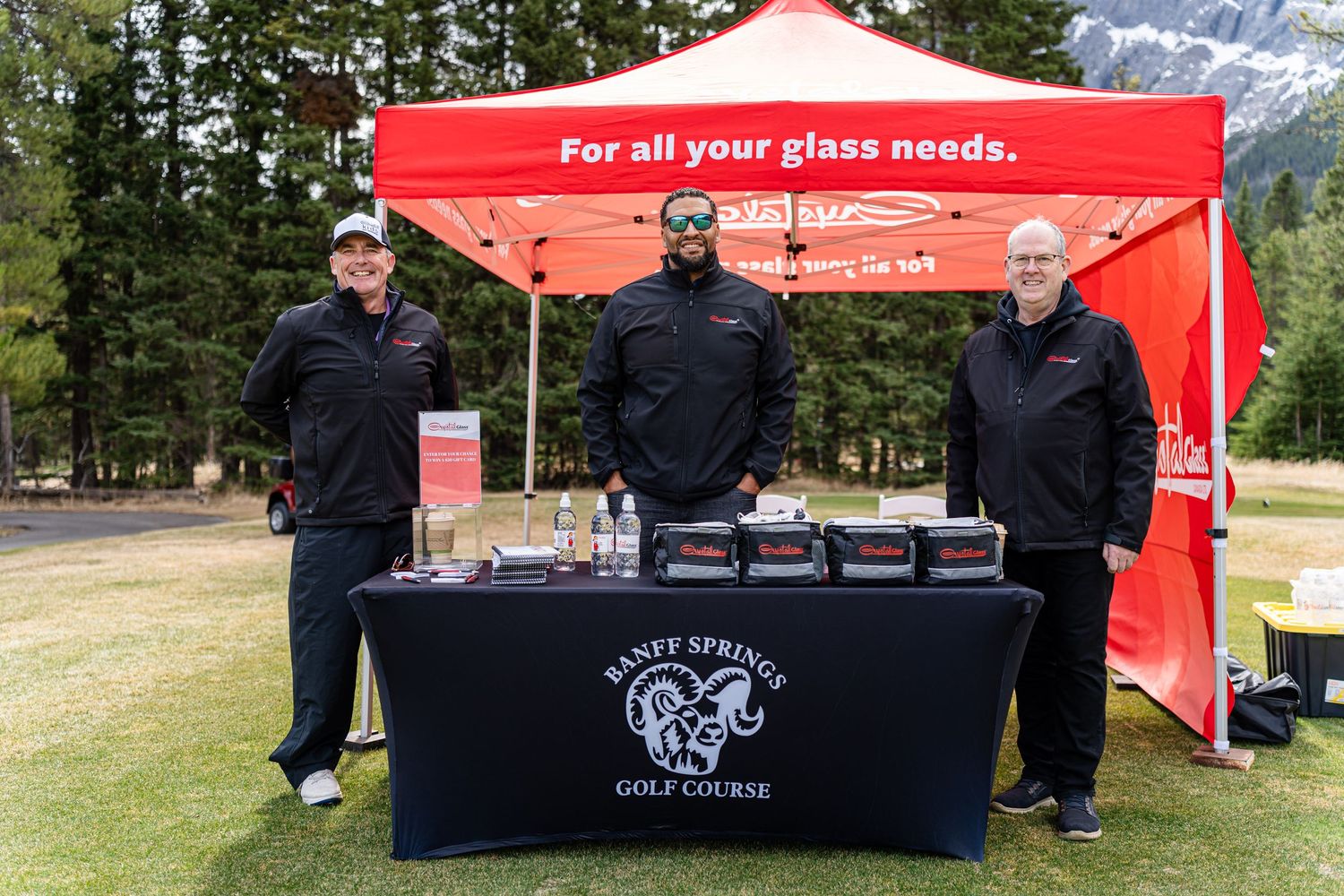 Three staff members stand behind a display table with a red tent at an outdoor event in a mountain setting.