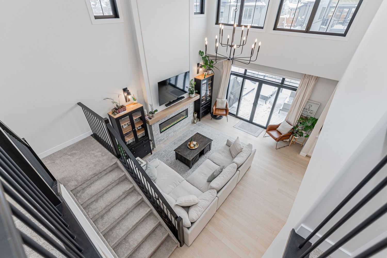 Aerial view of modern two-story living room with gray sofa, fireplace, chandelier, and floor-to-ceiling windows.