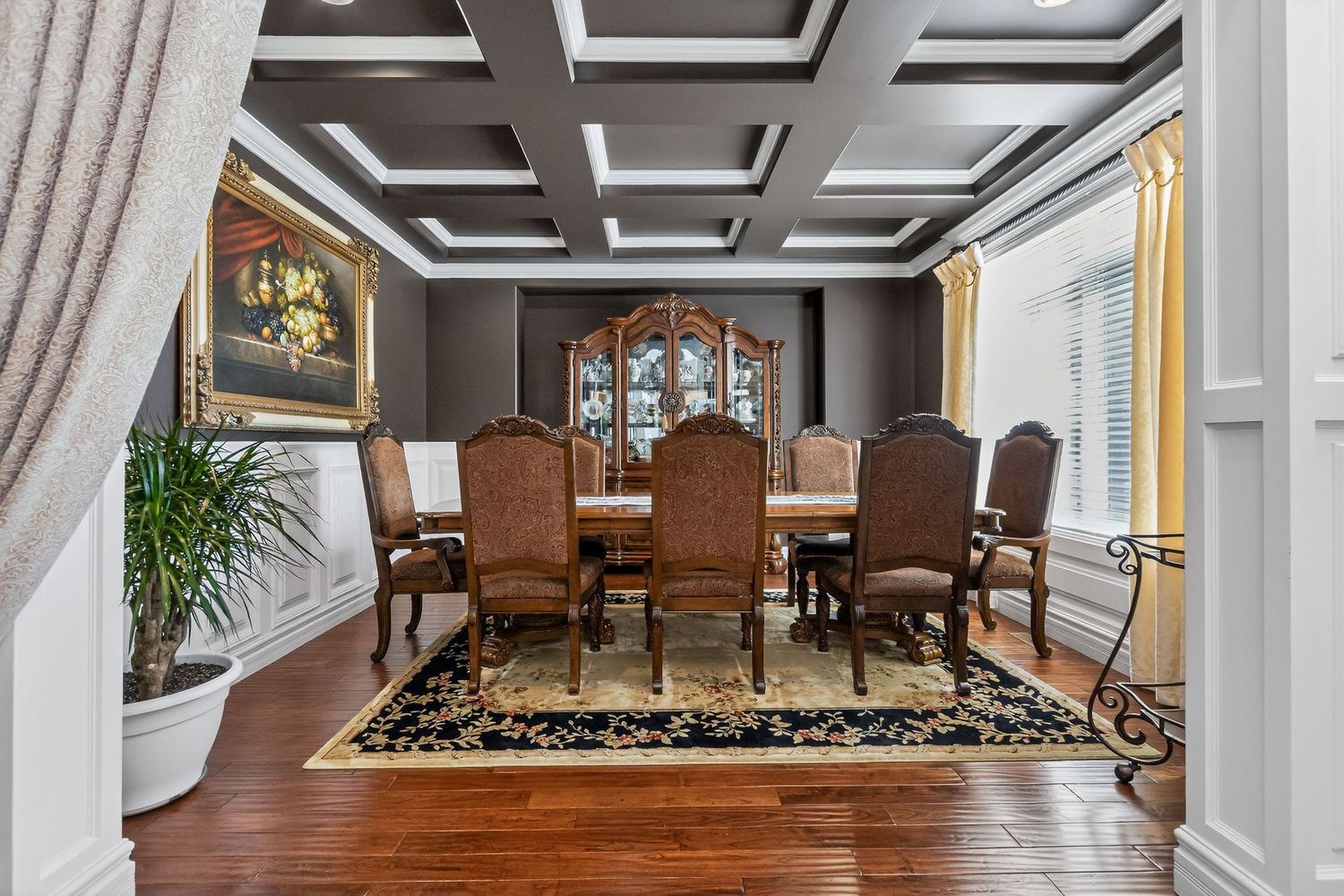 Elegant dining room with coffered ceiling, dark wood furniture, ornate china cabinet, and hardwood floors with floral rug.