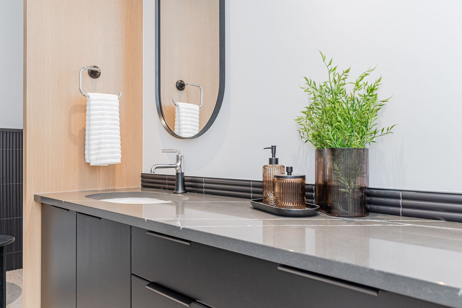 Modern bathroom vanity with dark cabinets, gray countertop, oval mirror, white towels, and green plant decor.