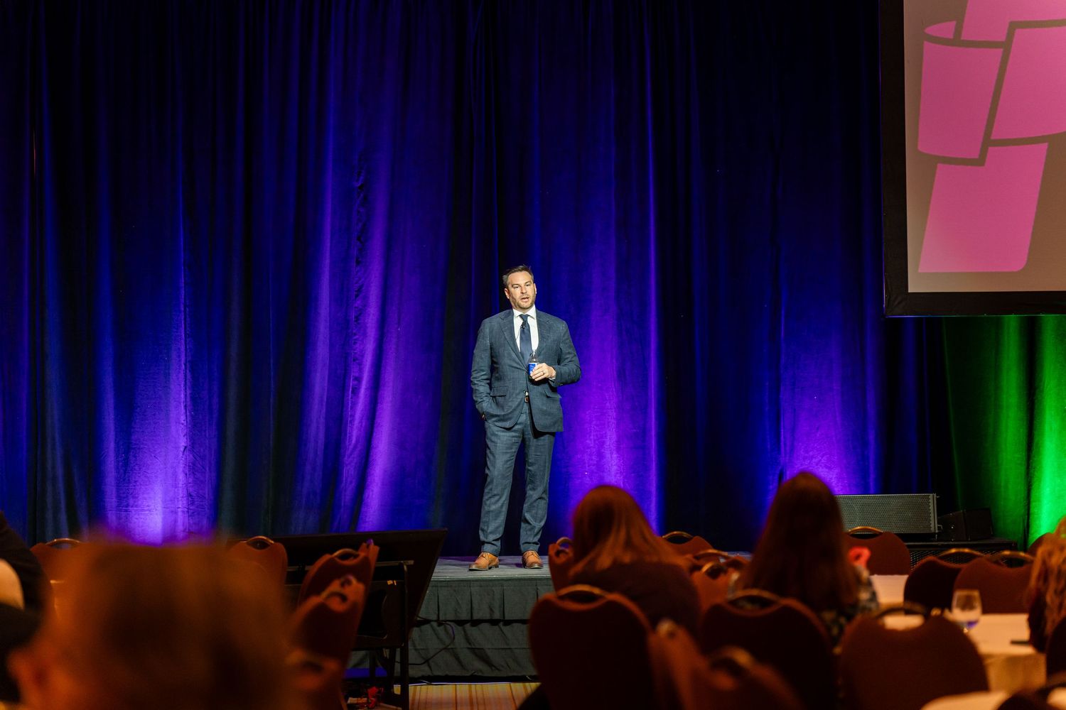 Presenter speaks on stage with colorful lighting and projection screen at conference venue.