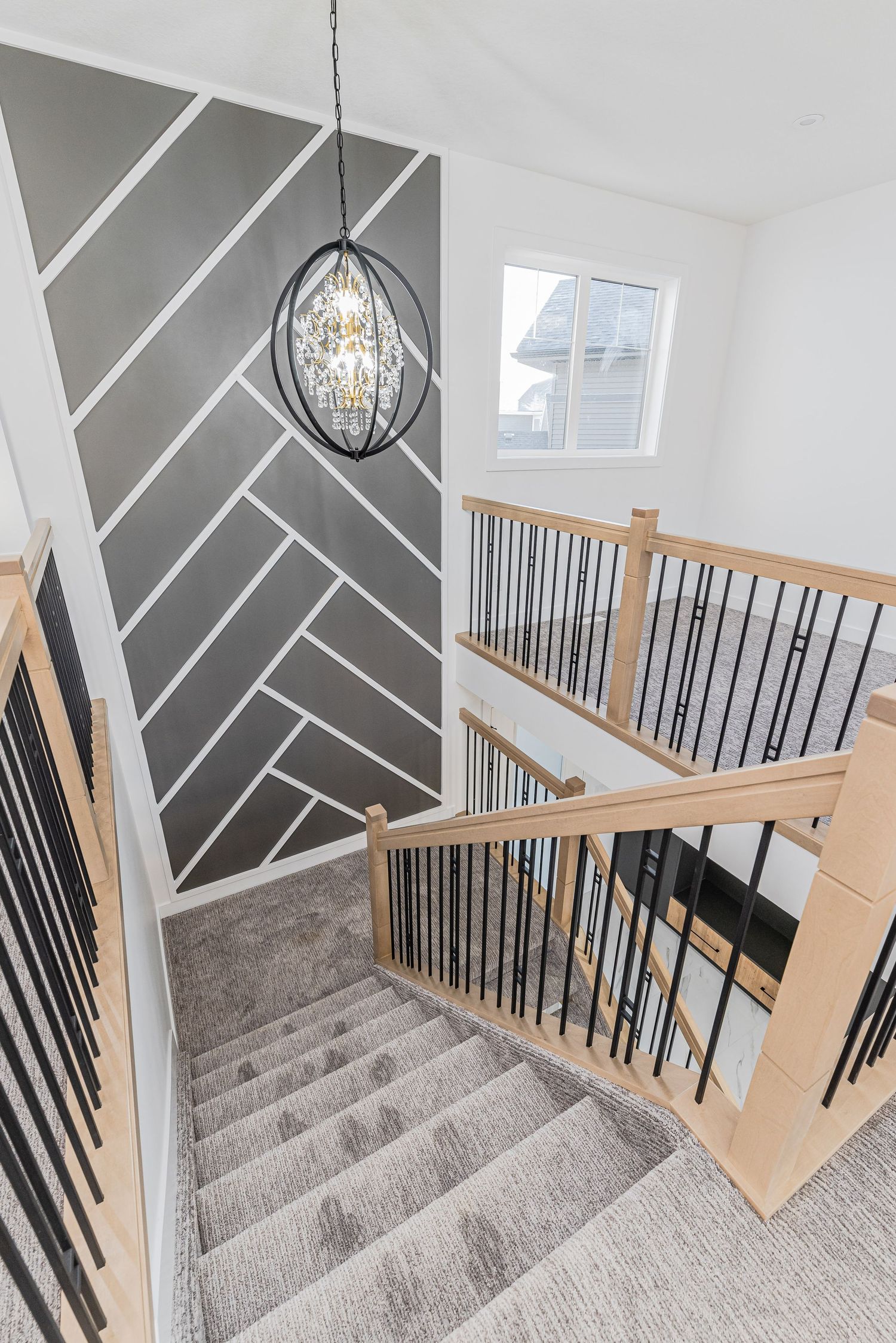 Modern staircase with gray carpet, wood and black iron railing, herringbone accent wall, and elegant pendant chandelier.