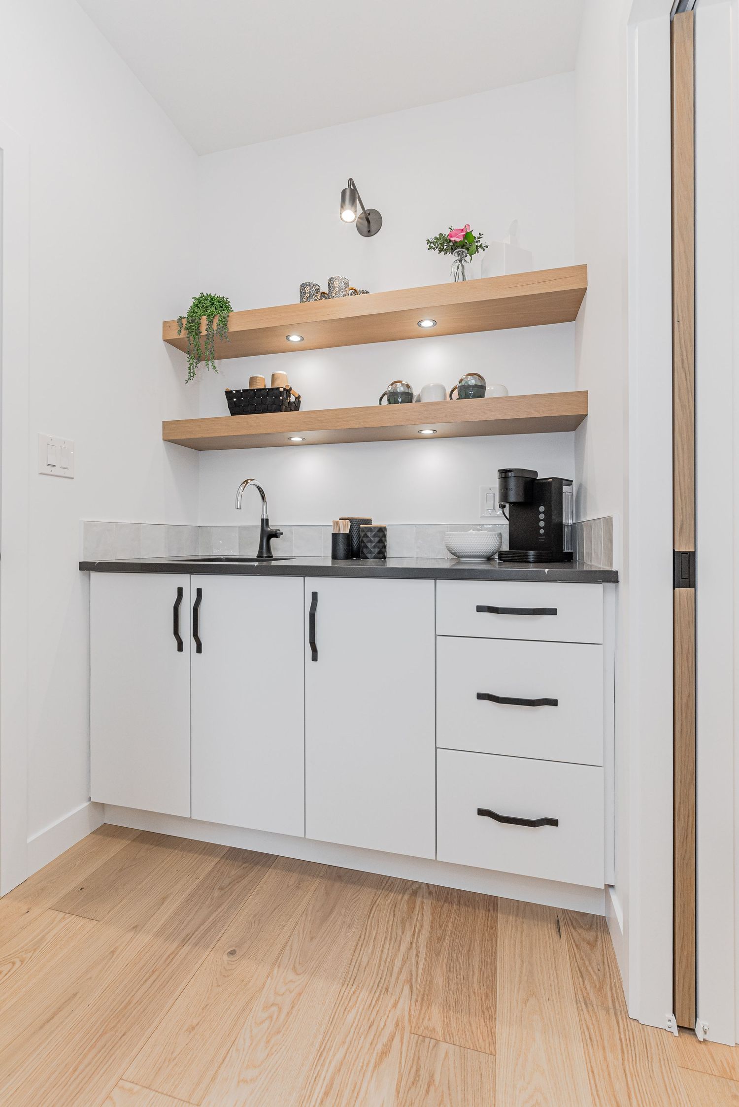 Modern white kitchen nook with open wood shelves, black countertop, sink, and coffee station on light hardwood floors.