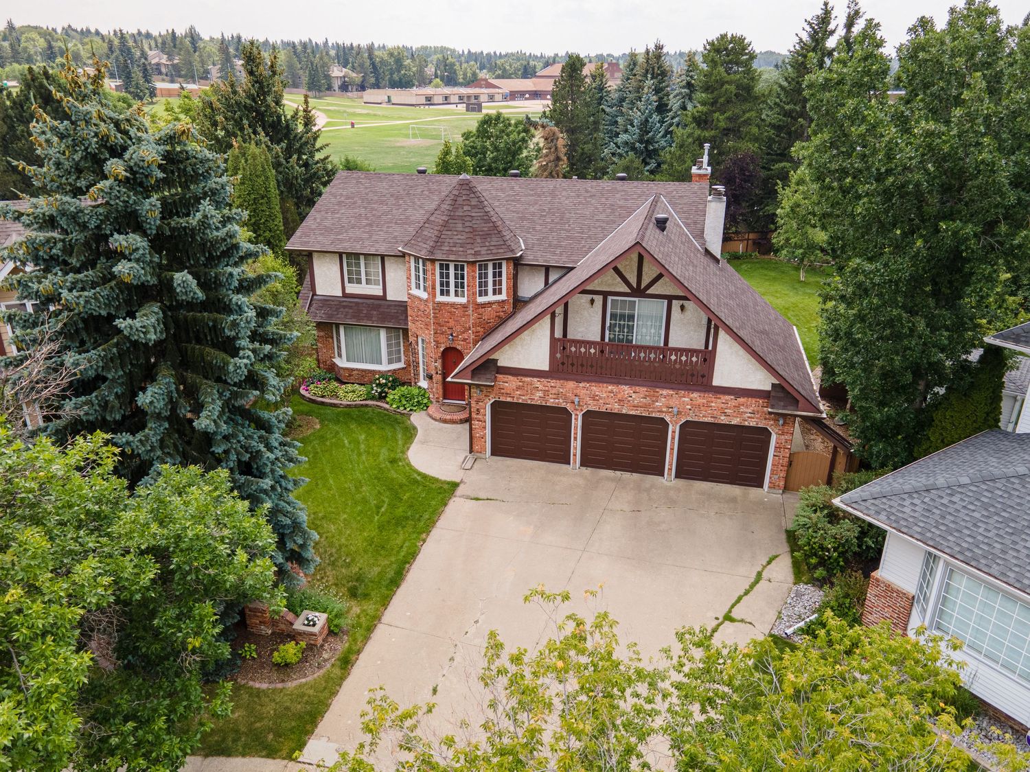 Aerial view of a Tudor-style brick home with triple garage, manicured lawn, and mature trees in a suburban neighborhood.