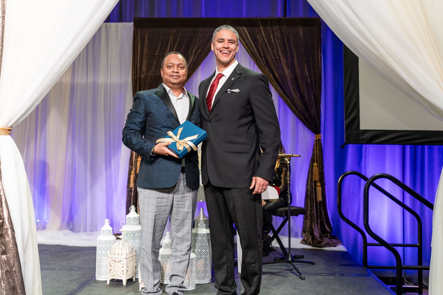 Two people pose with an award under white drapes at a formal event venue.