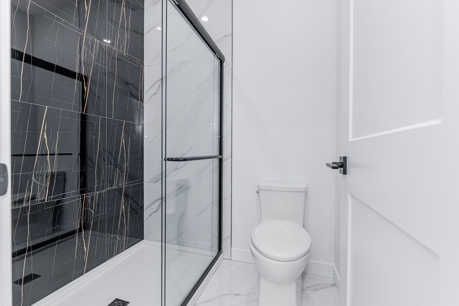 Modern bathroom with black marble walk-in shower, glass door, white toilet, and marble tile flooring.