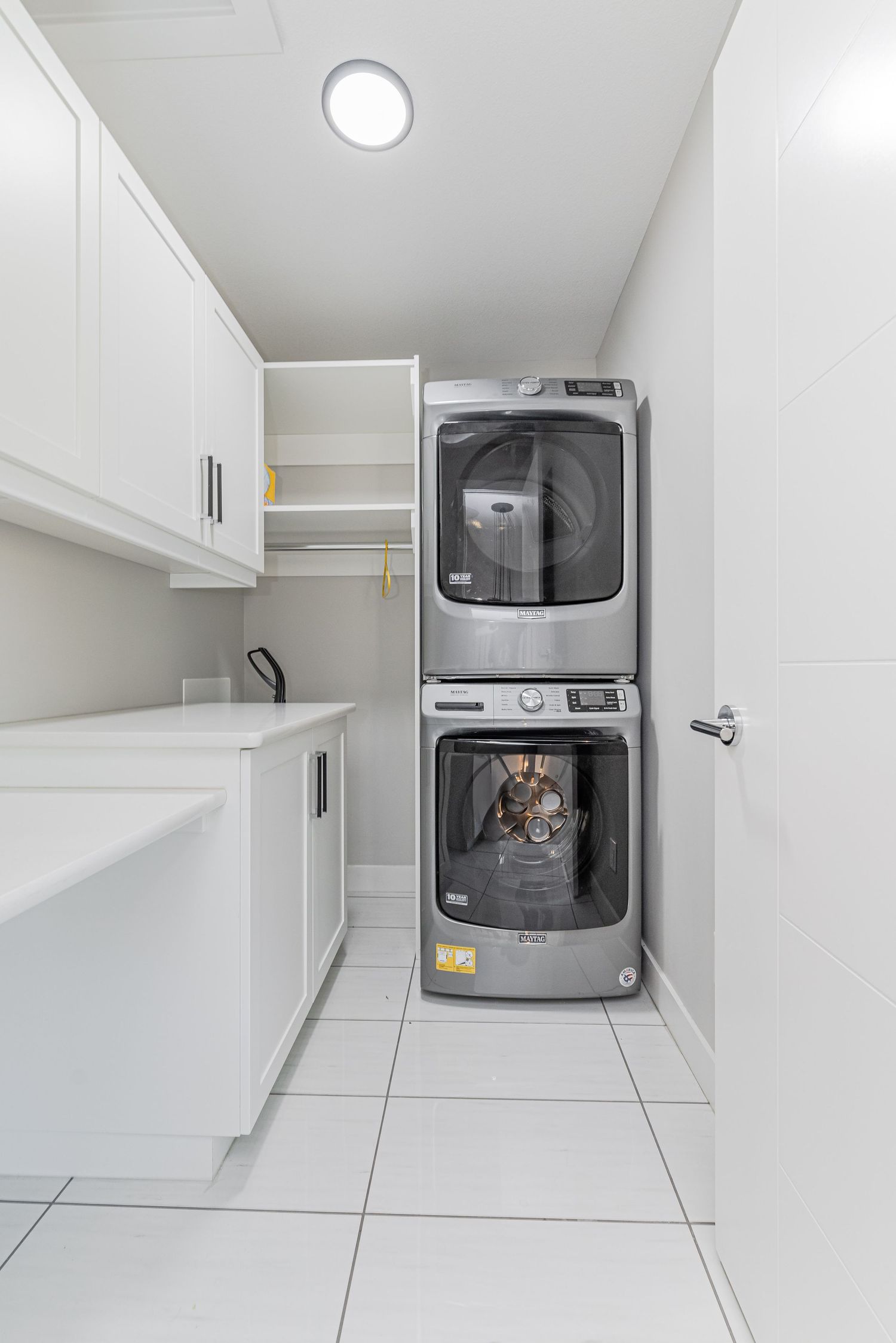 Modern laundry room with stacked silver washer and dryer, white cabinets, open shelving, and white tile flooring.