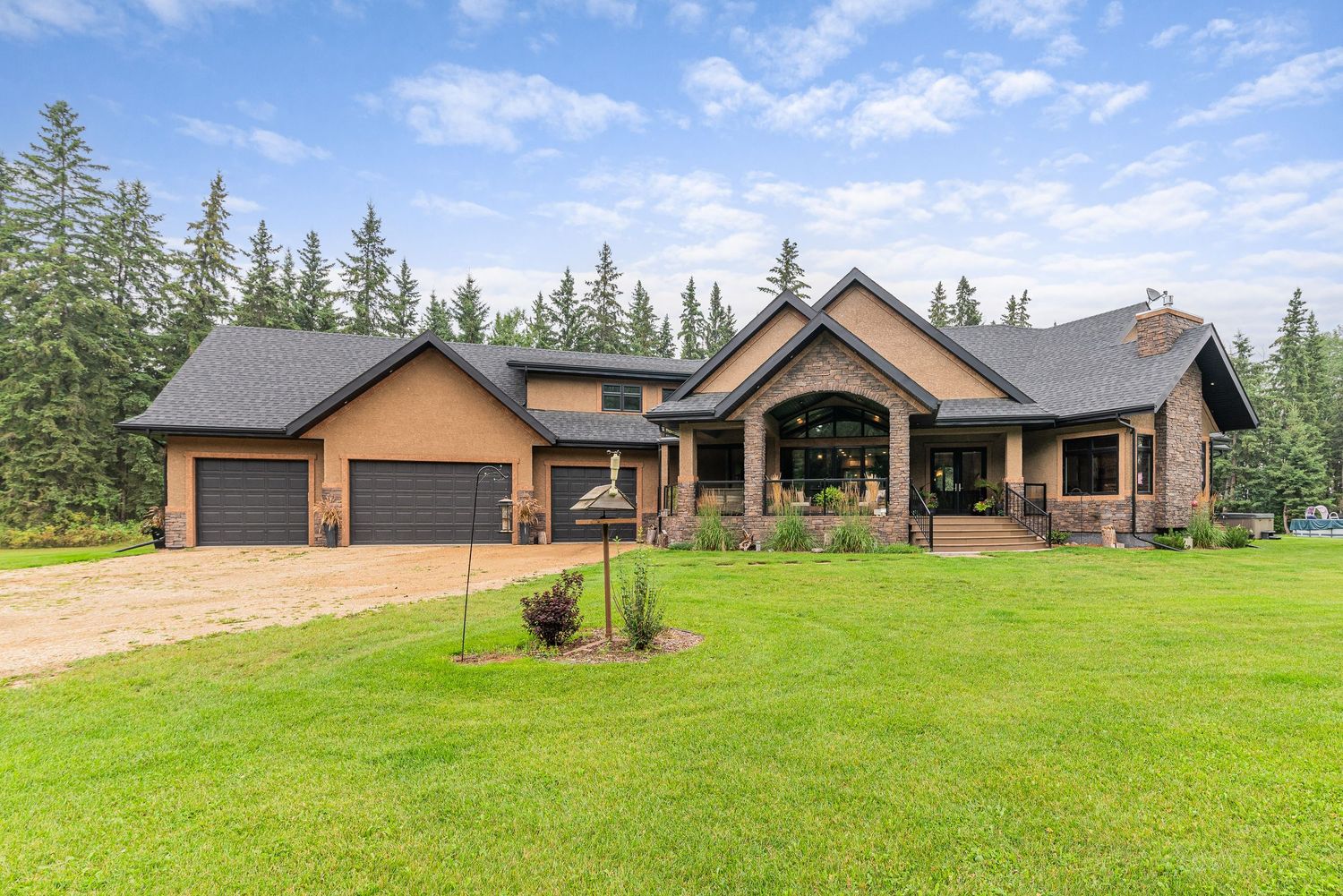 Luxury craftsman-style home with dark roof, stone accents, double garage, and manicured lawn surrounded by tall evergreen trees.