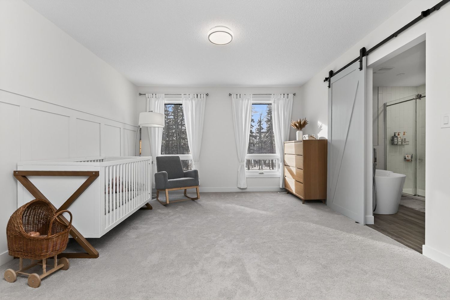 Bright nursery with white crib, barn door, carpet, and natural light from a large window.