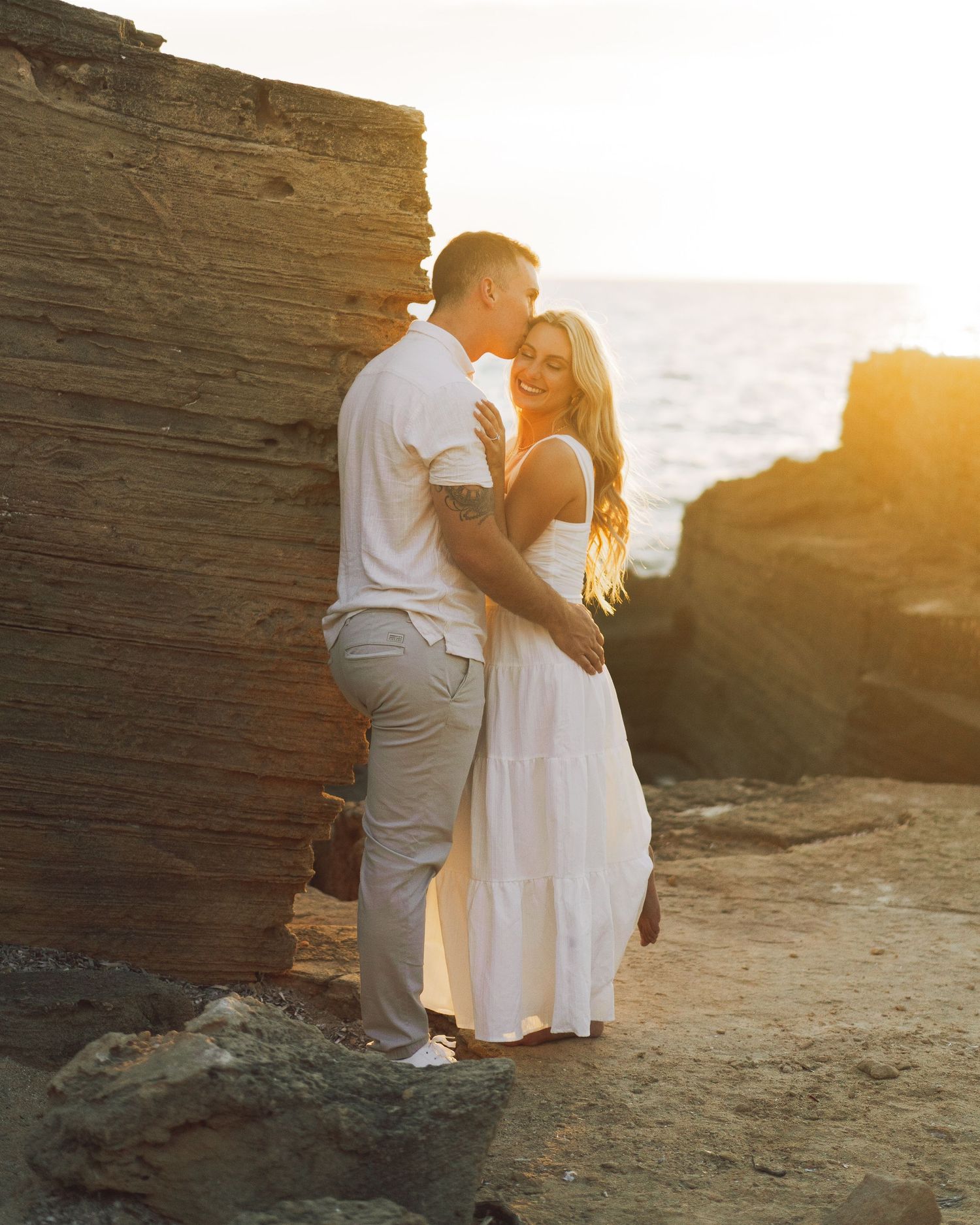 Couple in white outfits embracing on rocky beach cliffs during golden hour.