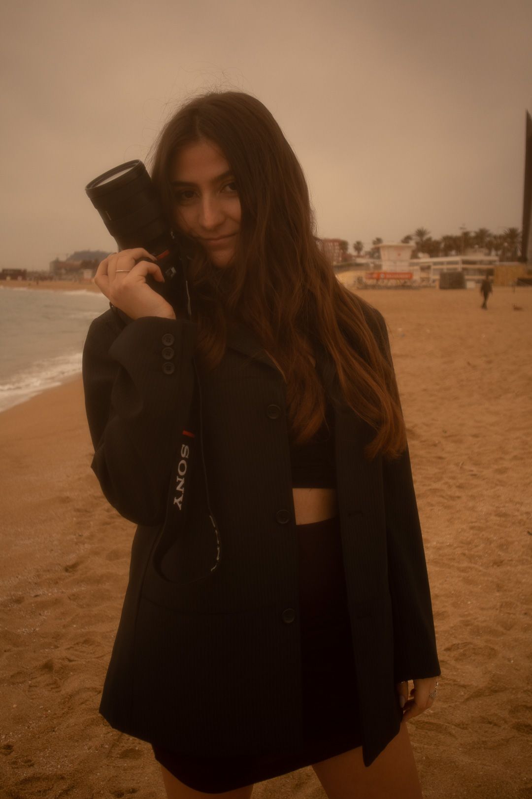 A person in dark clothing stands on a moody, sepia-toned beach during sunset.