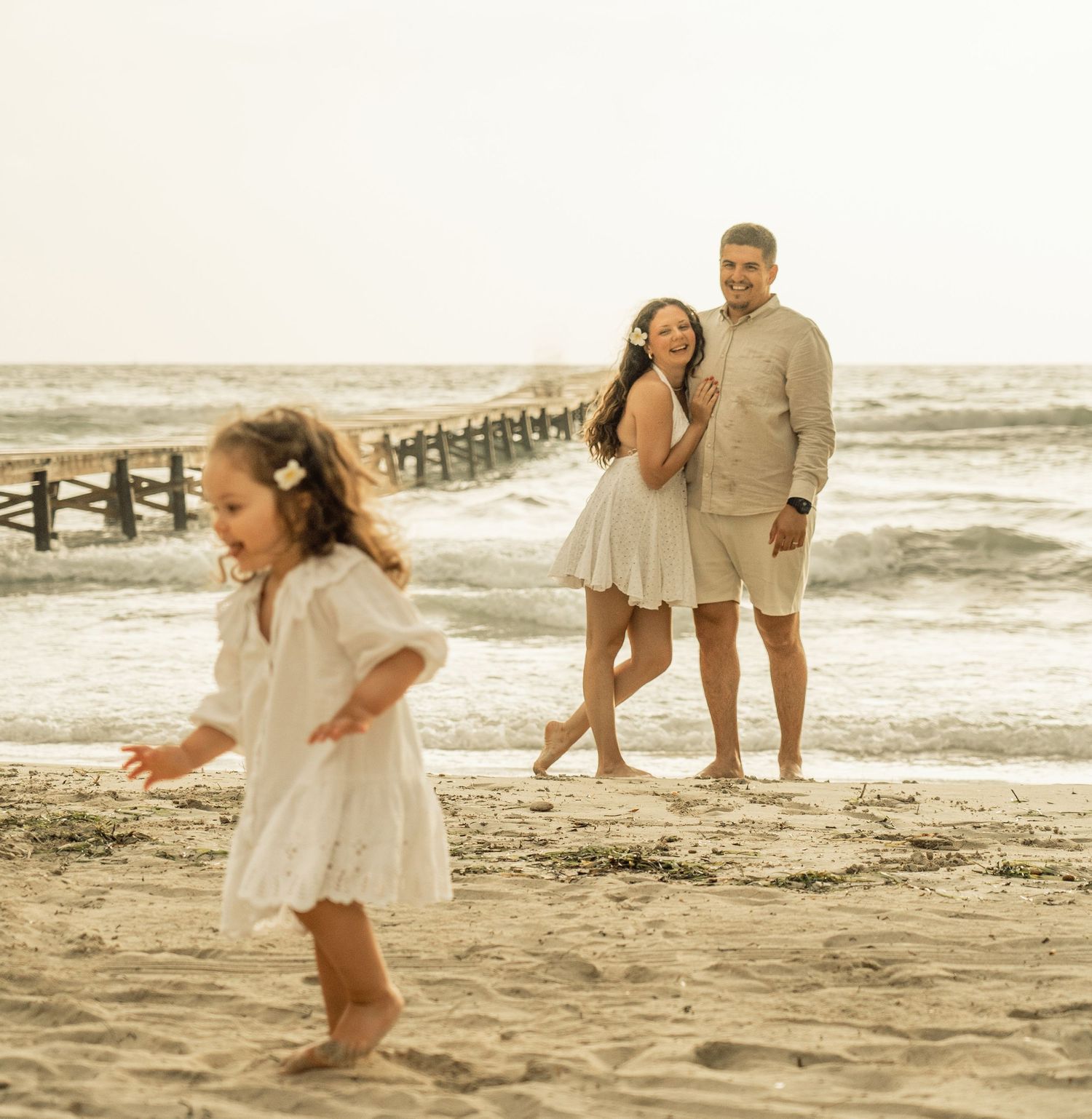 Family enjoying a sunset walk along a sandy beach with waves crashing against a wooden pier in the background.