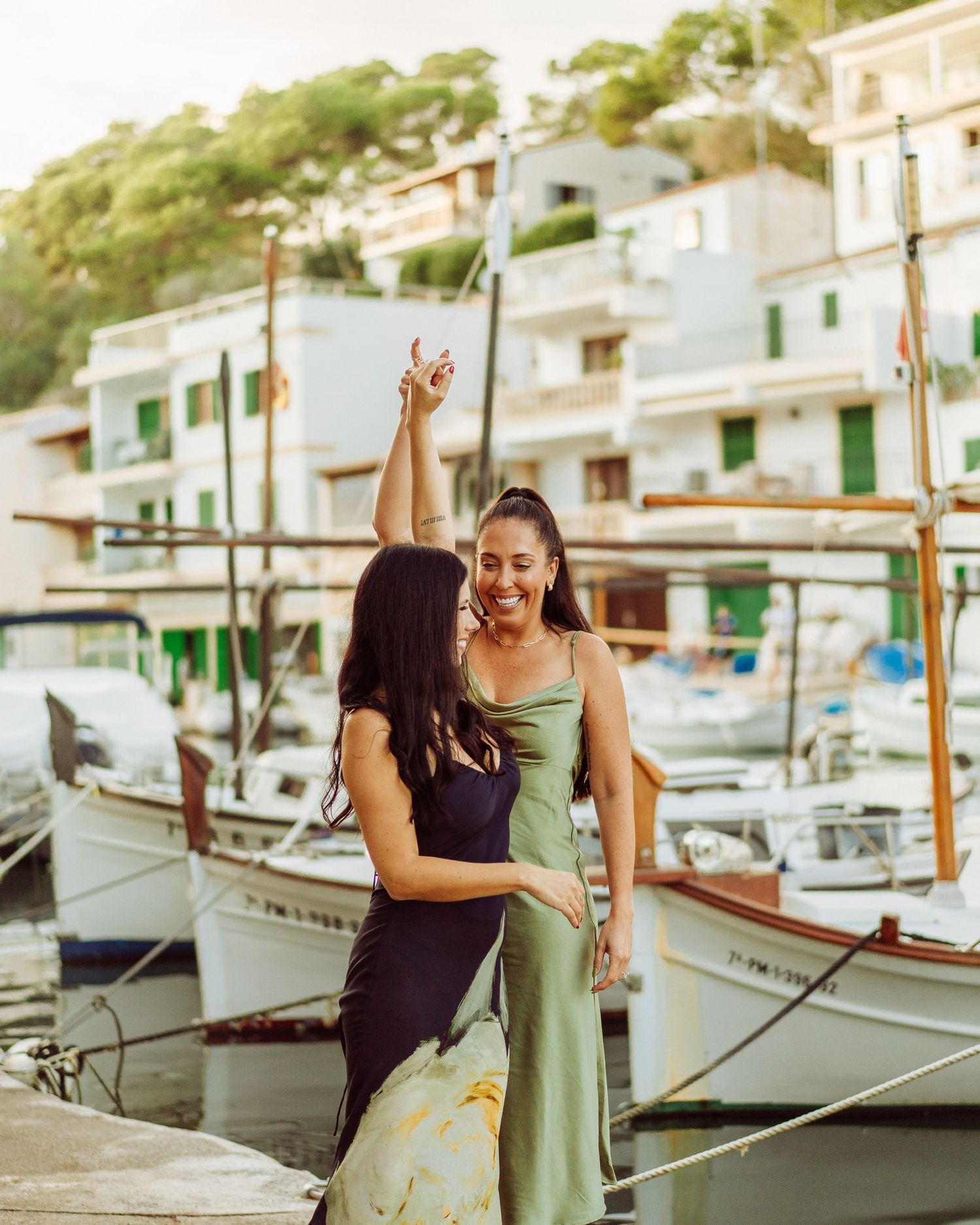 Two people laughing together on a dock with boats moored in the background during sunset at a Mediterranean harbor.