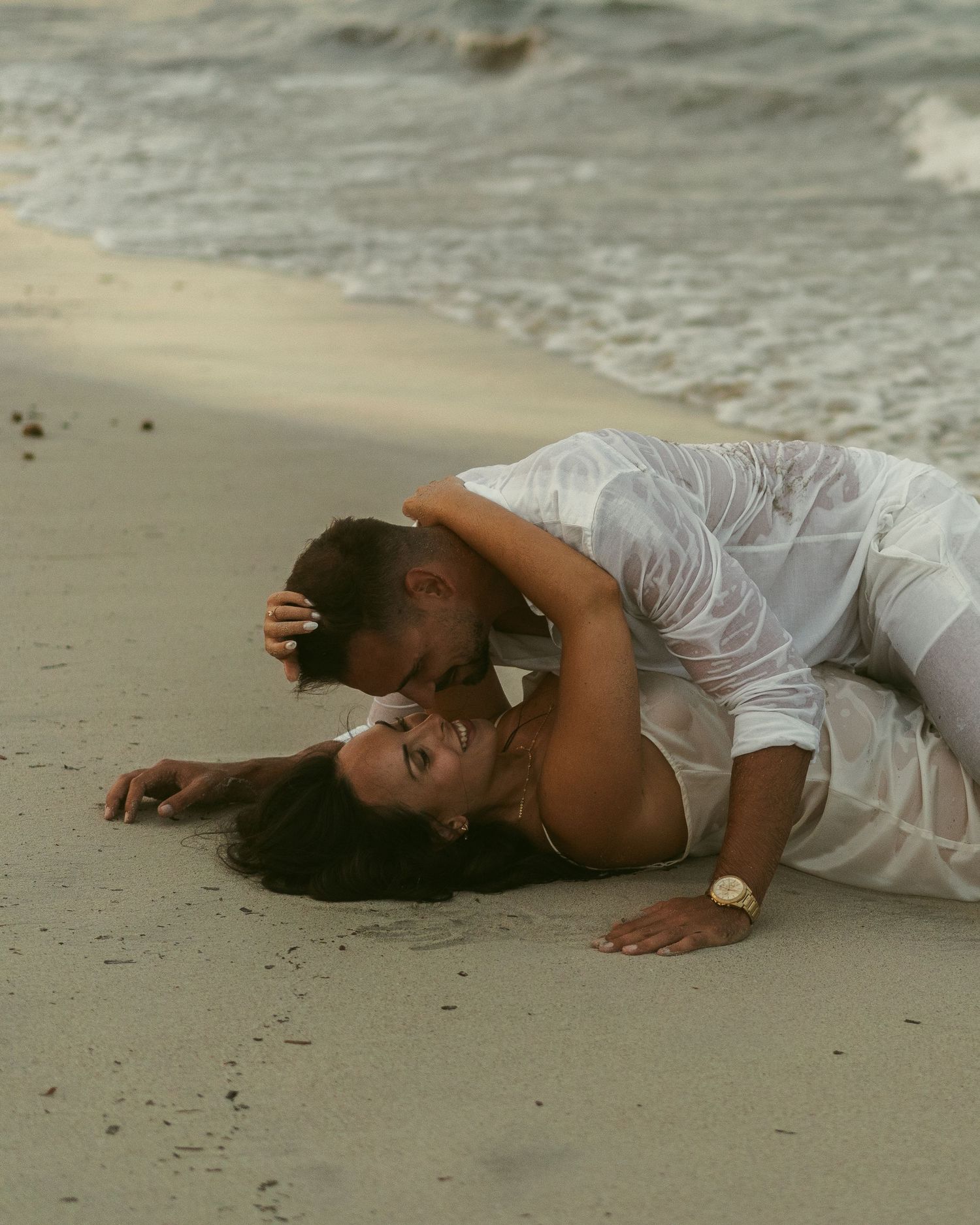 A person in a white dress lying on the sandy beach with waves in the background.