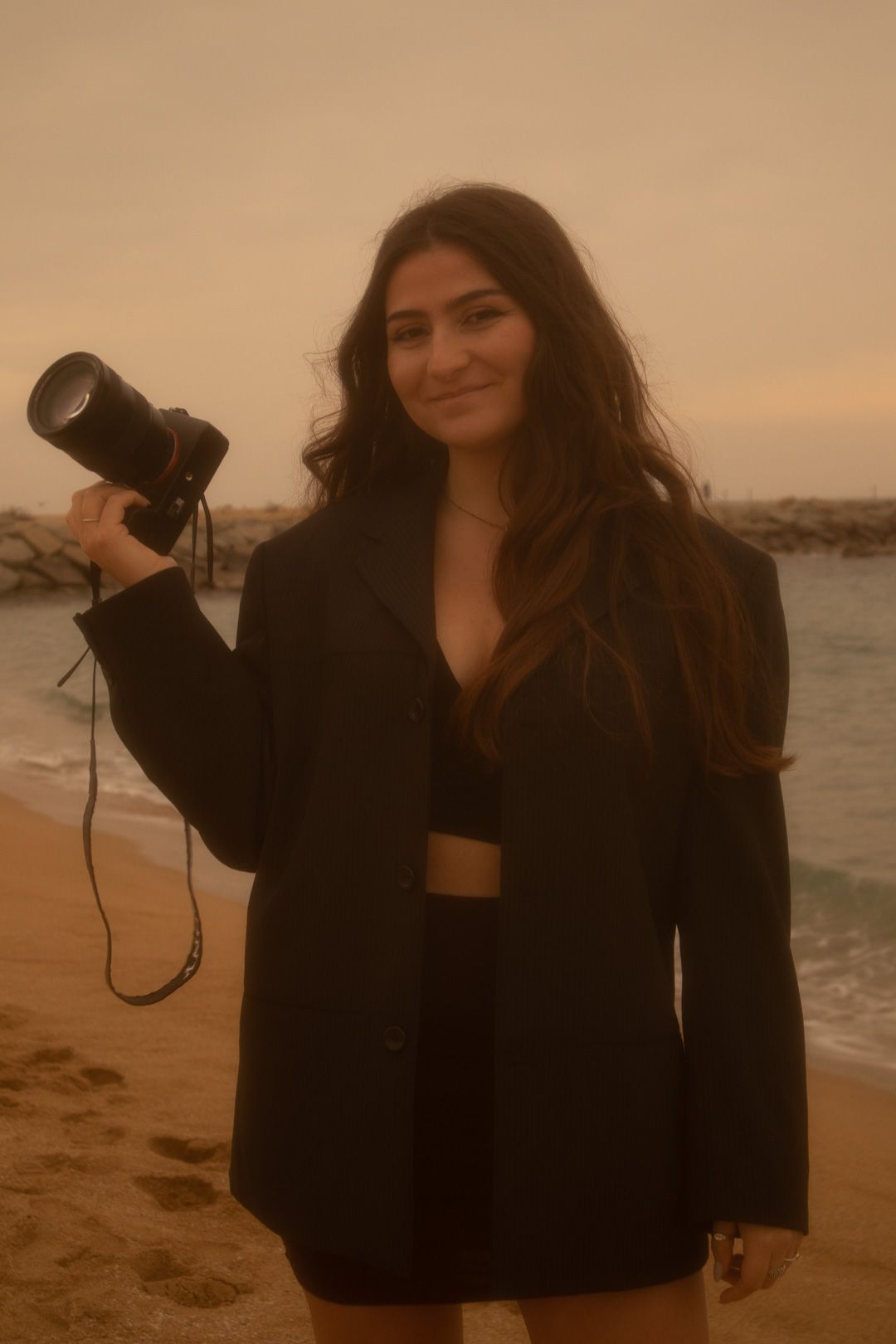 A person in a black jacket poses with a camera on a beach at sunset in a series of artistic photos.