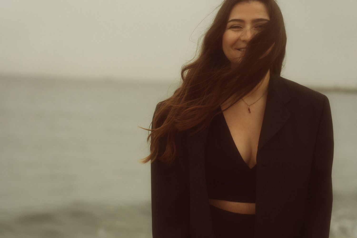 Professional fashion photoshoot on a beach with model wearing a black blazer and dress against moody coastal backdrop.
