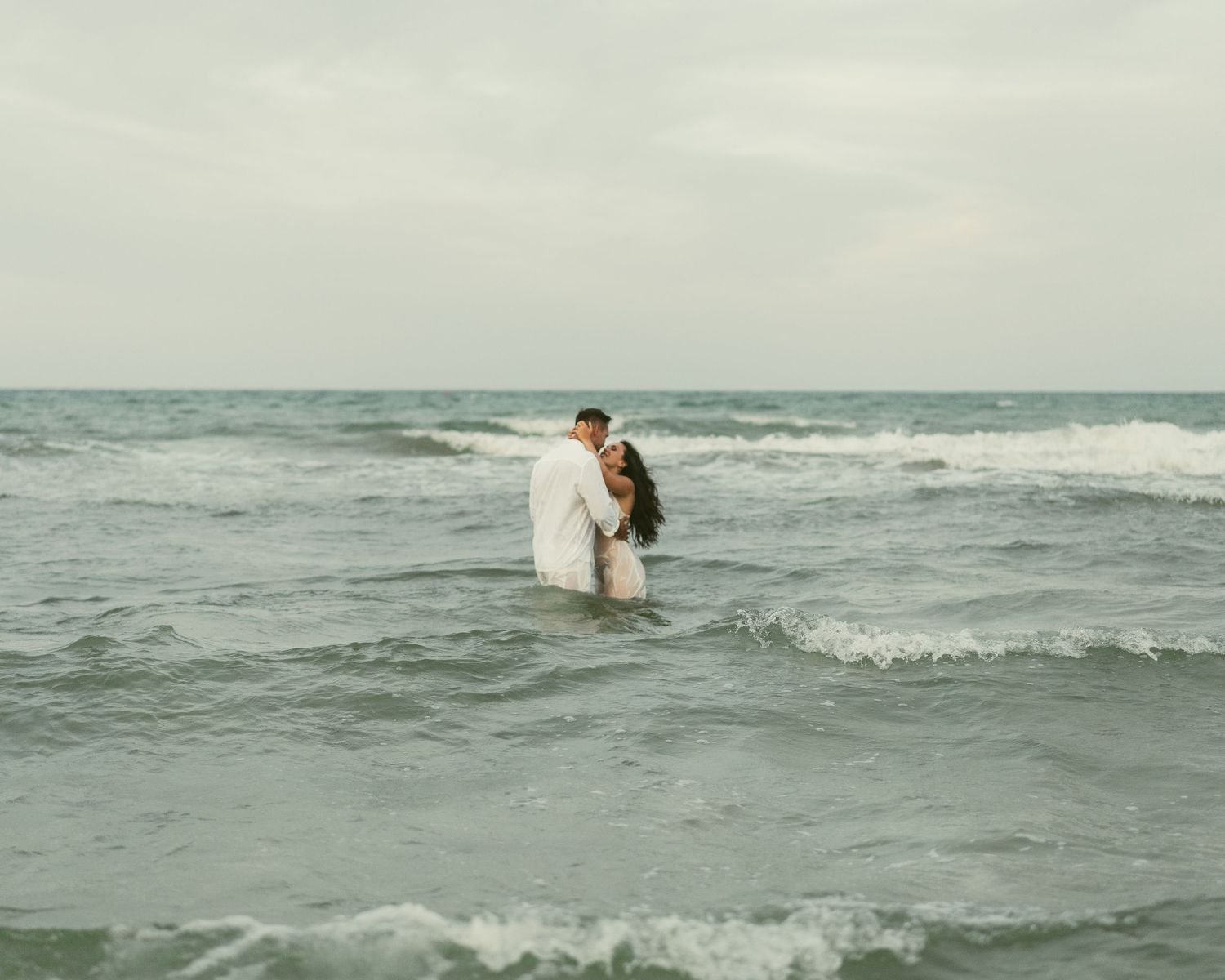 A couple embraces while standing in ocean waves during an overcast beach photoshoot.