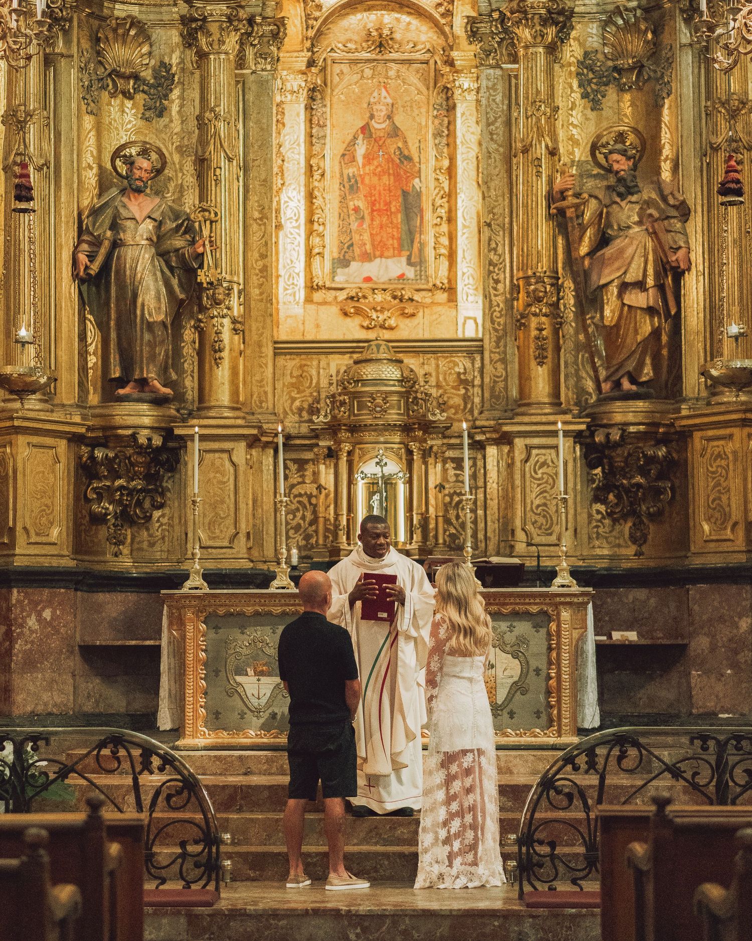 Catholic priest conducts religious ceremony at ornate baroque altar with golden decorations and religious statues.