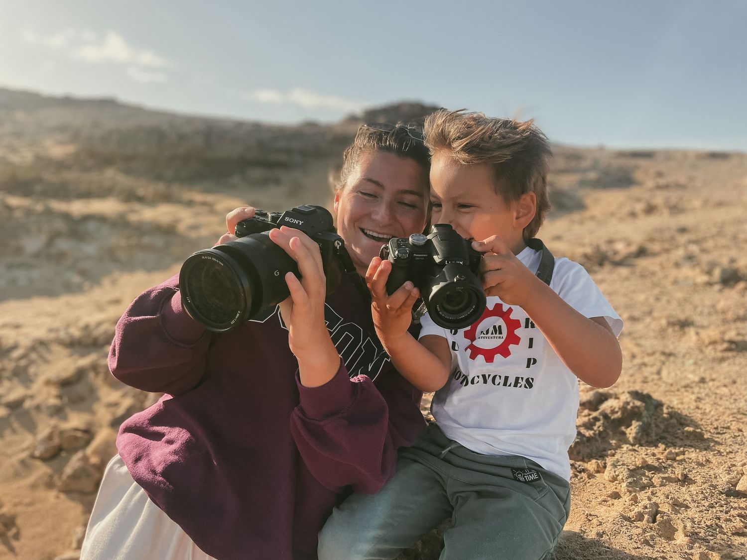 Two people share an intimate moment while taking photos in a desert landscape at sunset.