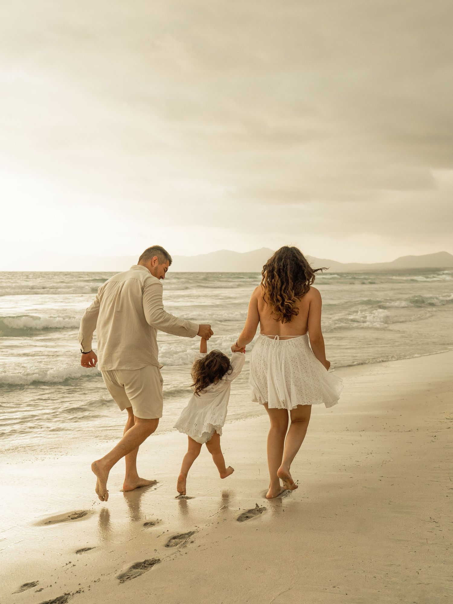 Three people walking along a beach at sunset leave footprints in the sand as waves crash on the shore.