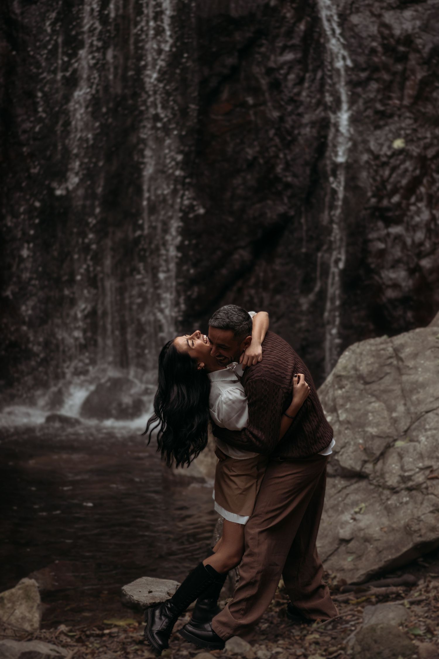 Two people share an intimate moment in front of a misty waterfall in a moody forest setting.