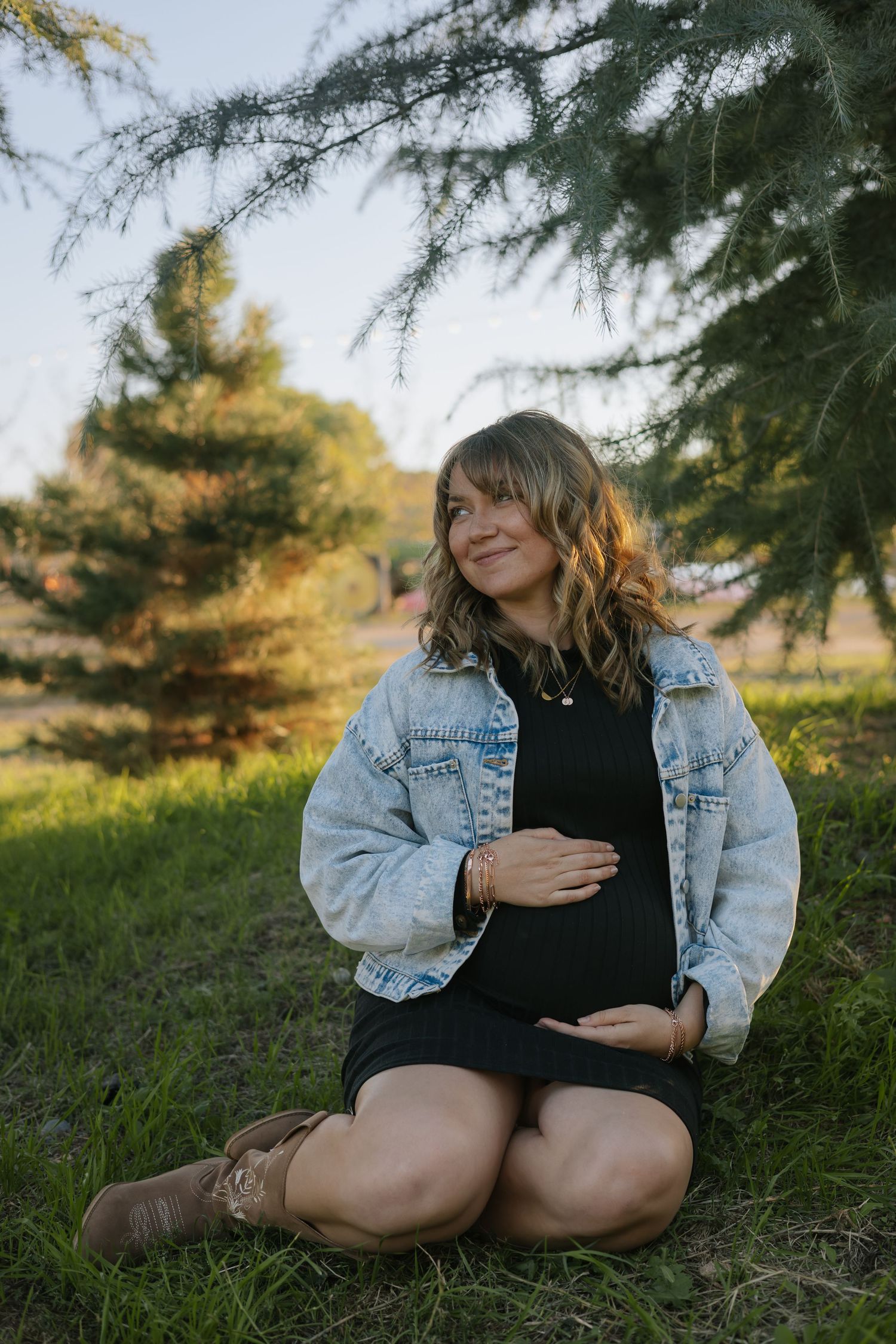 Expectant mother in denim jacket sitting on grass near evergreen trees.