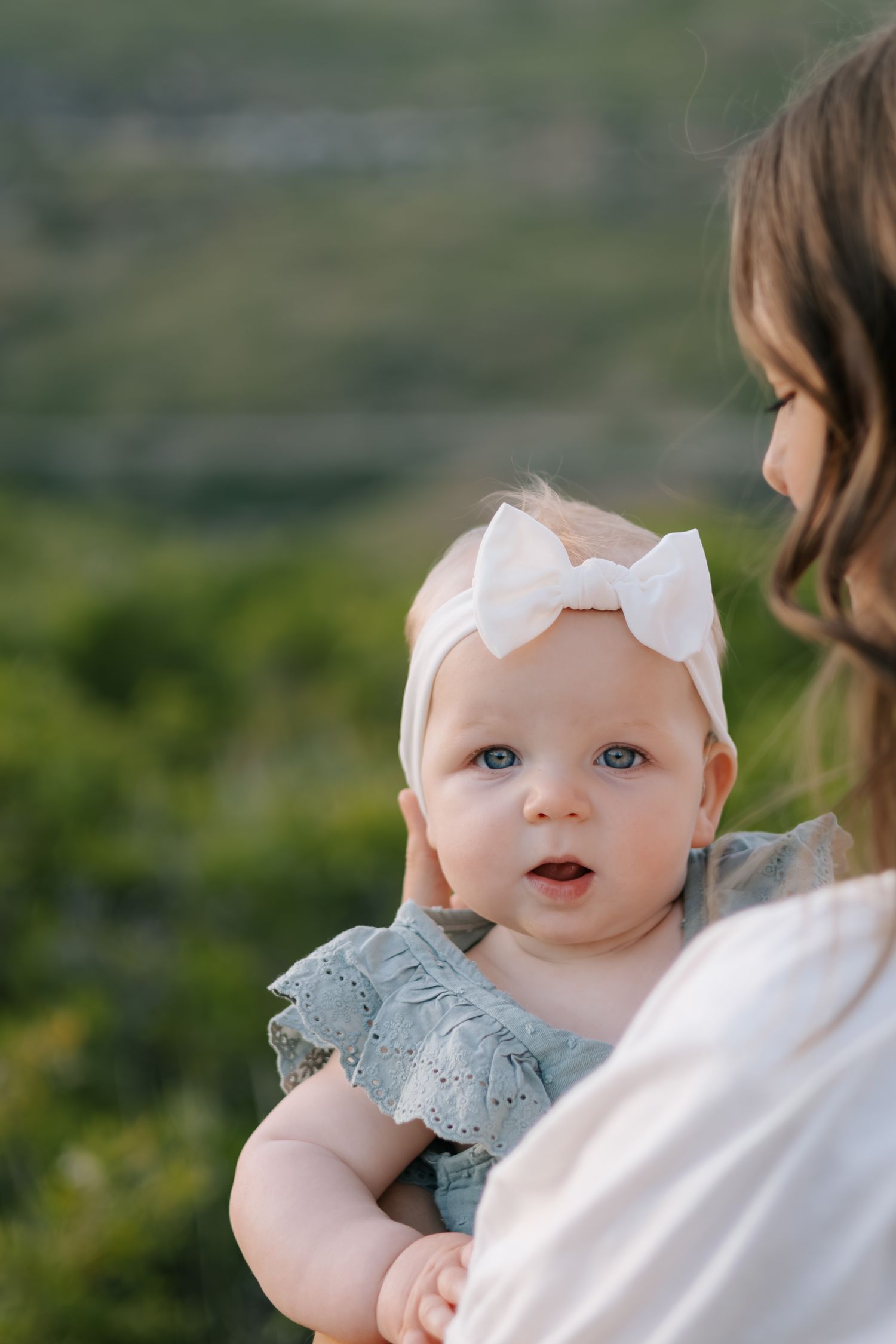 A baby in a blue dress and white bow headband looks over their shoulder outdoors.