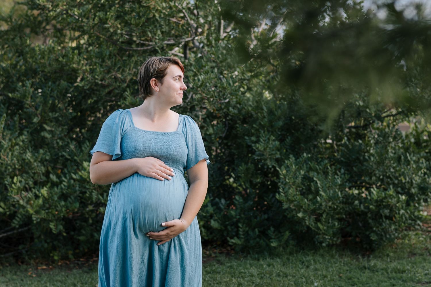 Maternity photoshoot in a light blue dress outdoors among greenery.