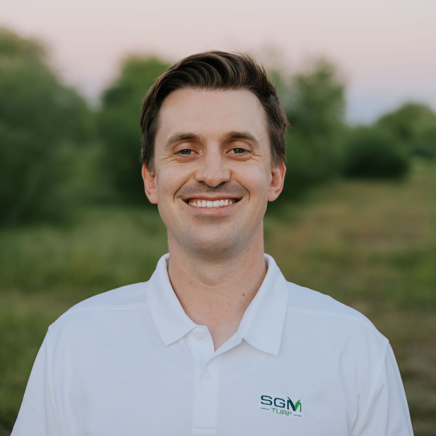 Person wearing white branded polo shirt smiling against sunset background.