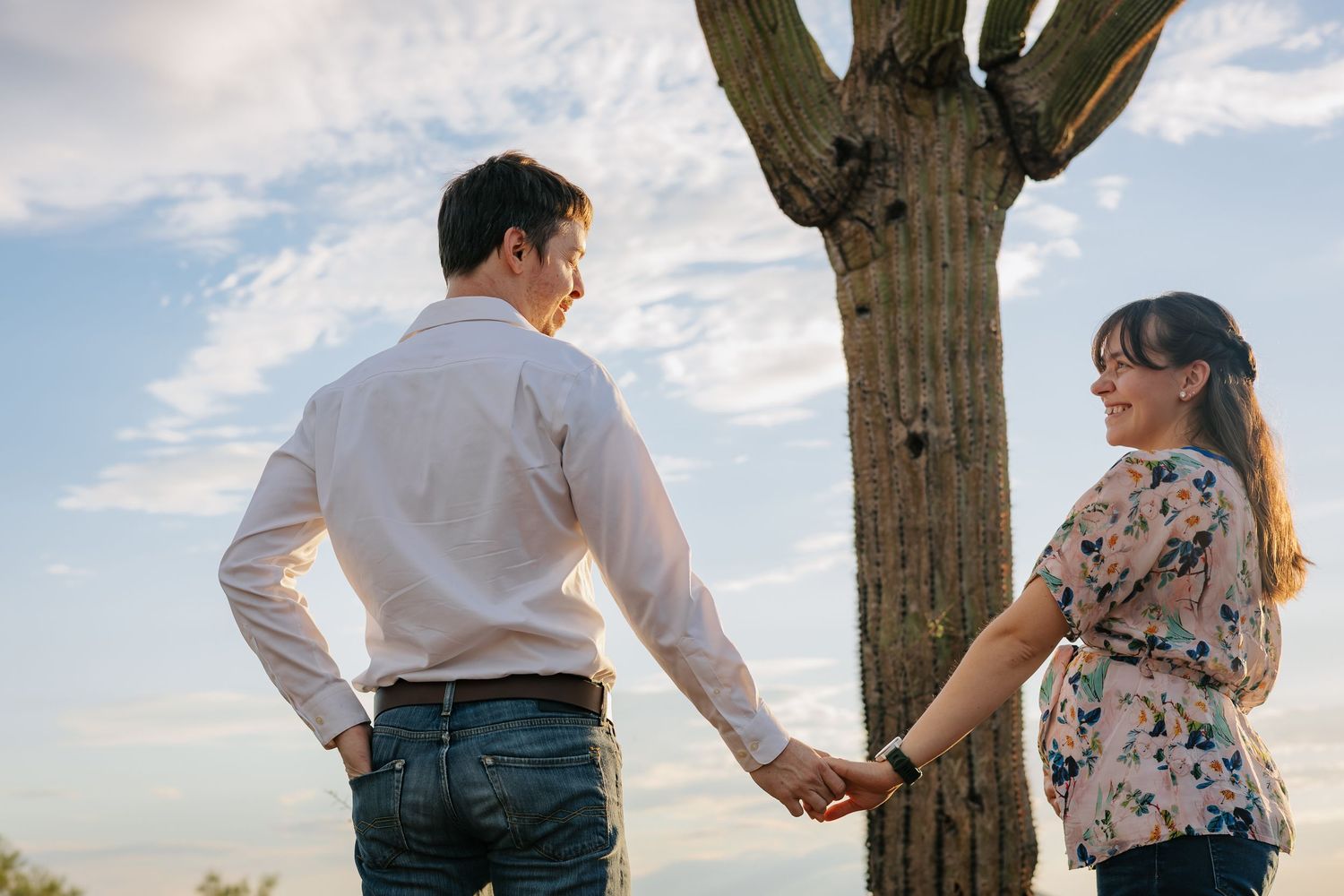 Couple holding hands in front of large saguaro cactus at sunset.