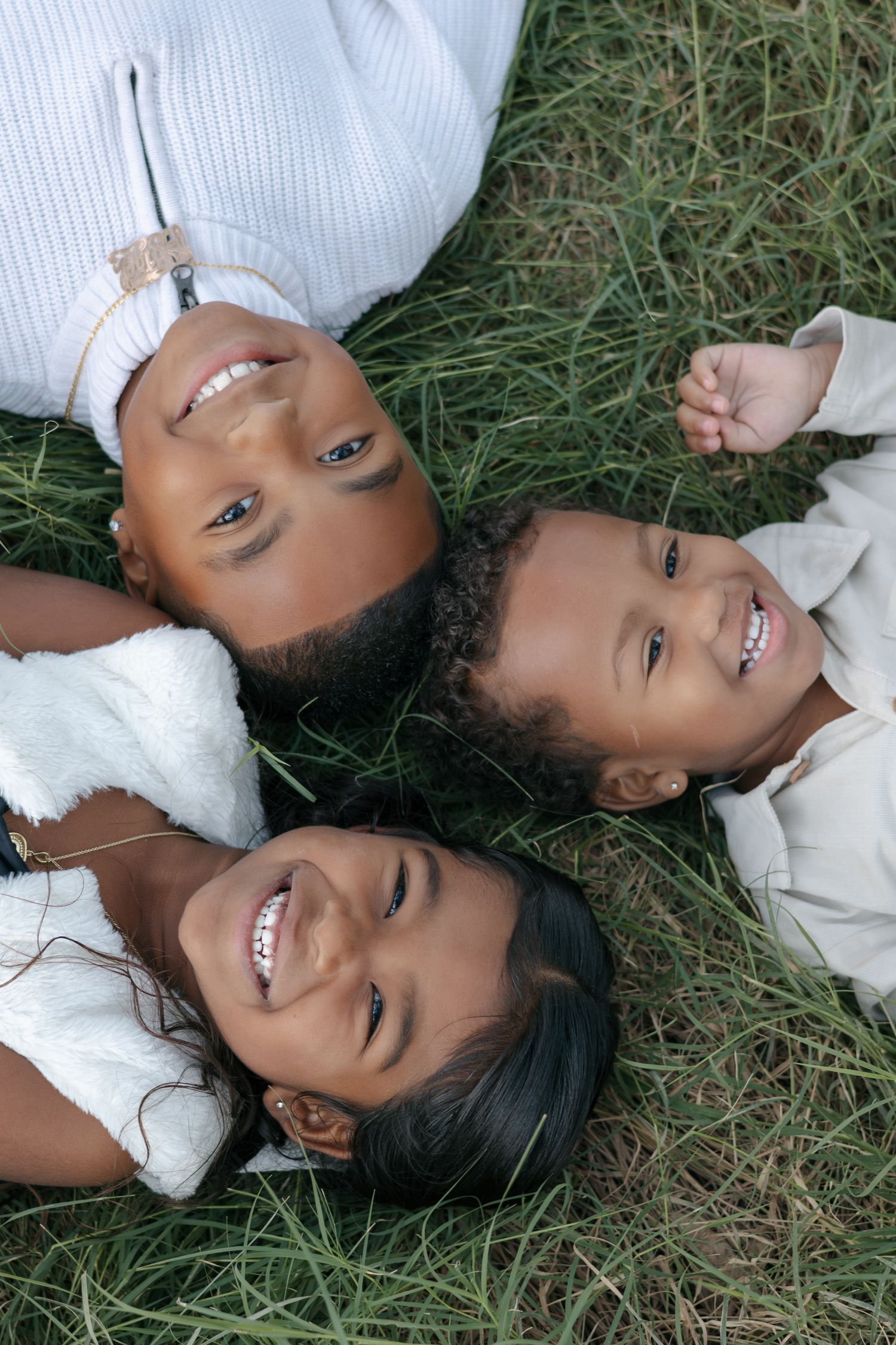 Three children lying on grass wearing white clothing while smiling at camera.