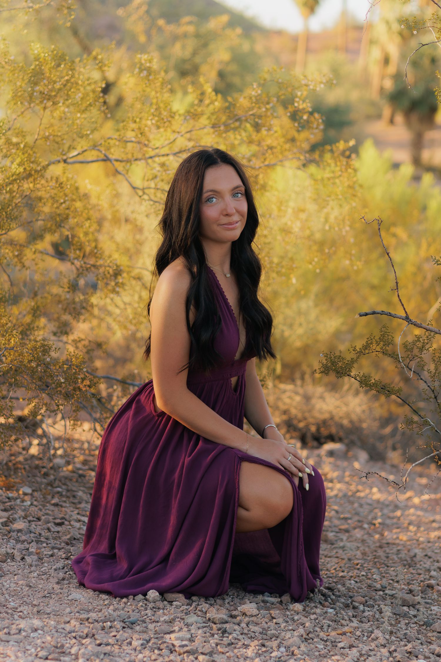Young woman in burgundy dress kneeling outdoors among desert shrubs in golden hour light.