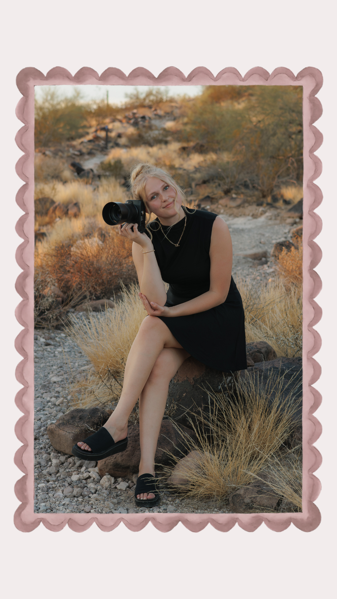 Blonde woman in black dress sitting on rocks in desert landscape holding a professional camera, smiling.
