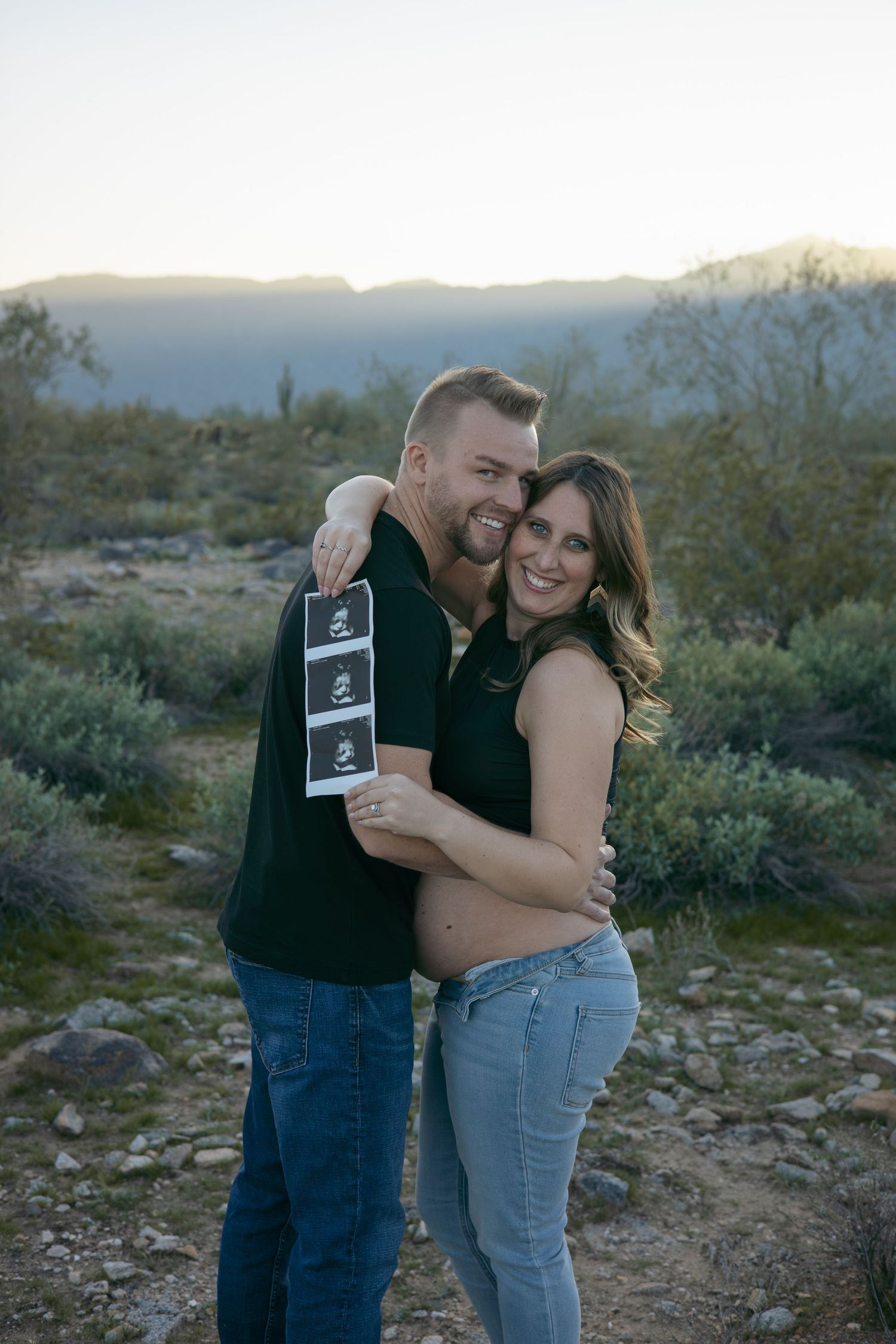 Couple holding ultrasound photos in a desert landscape at sunset.