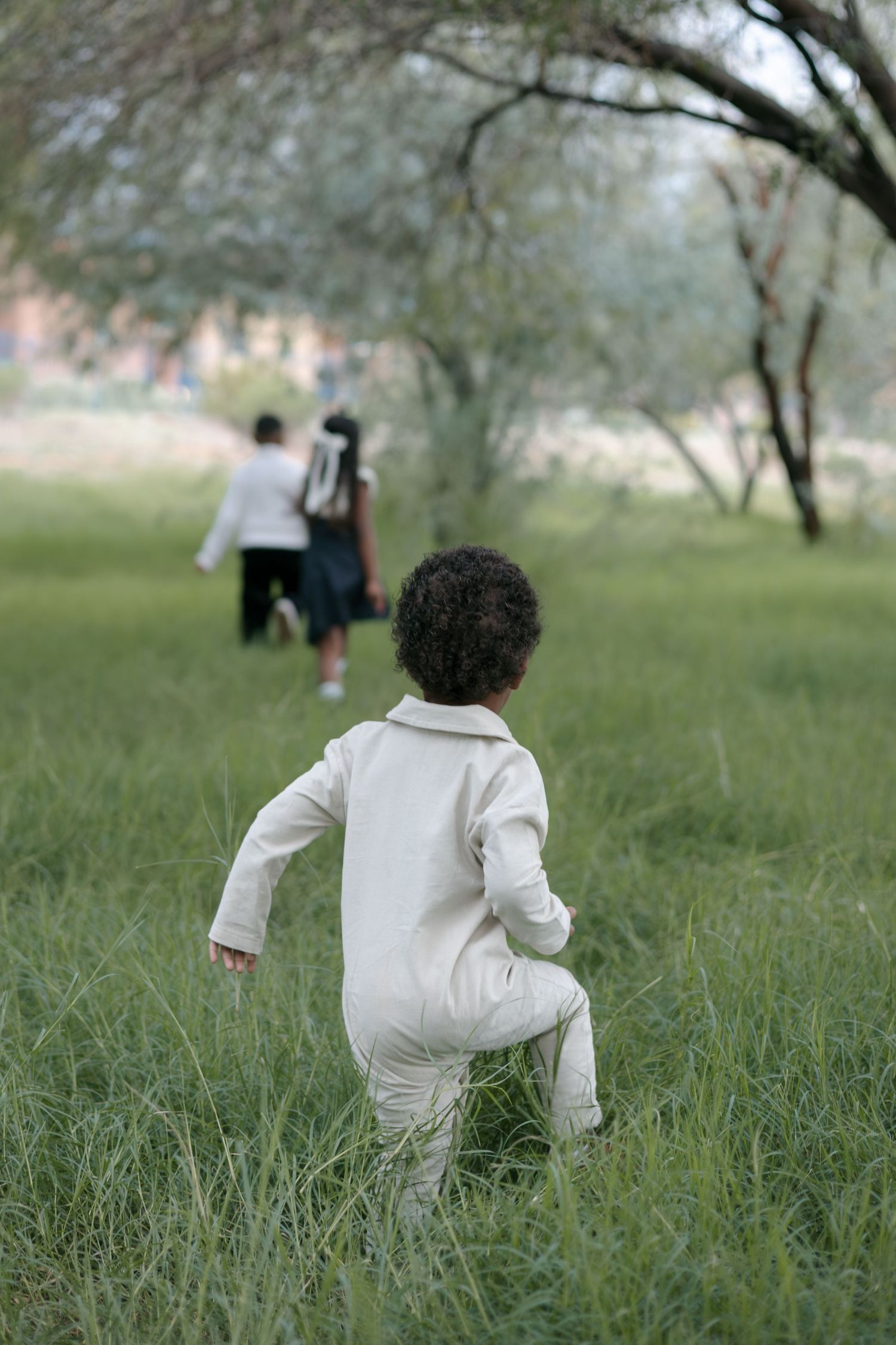Child in white outfit running through tall grass with others in background.