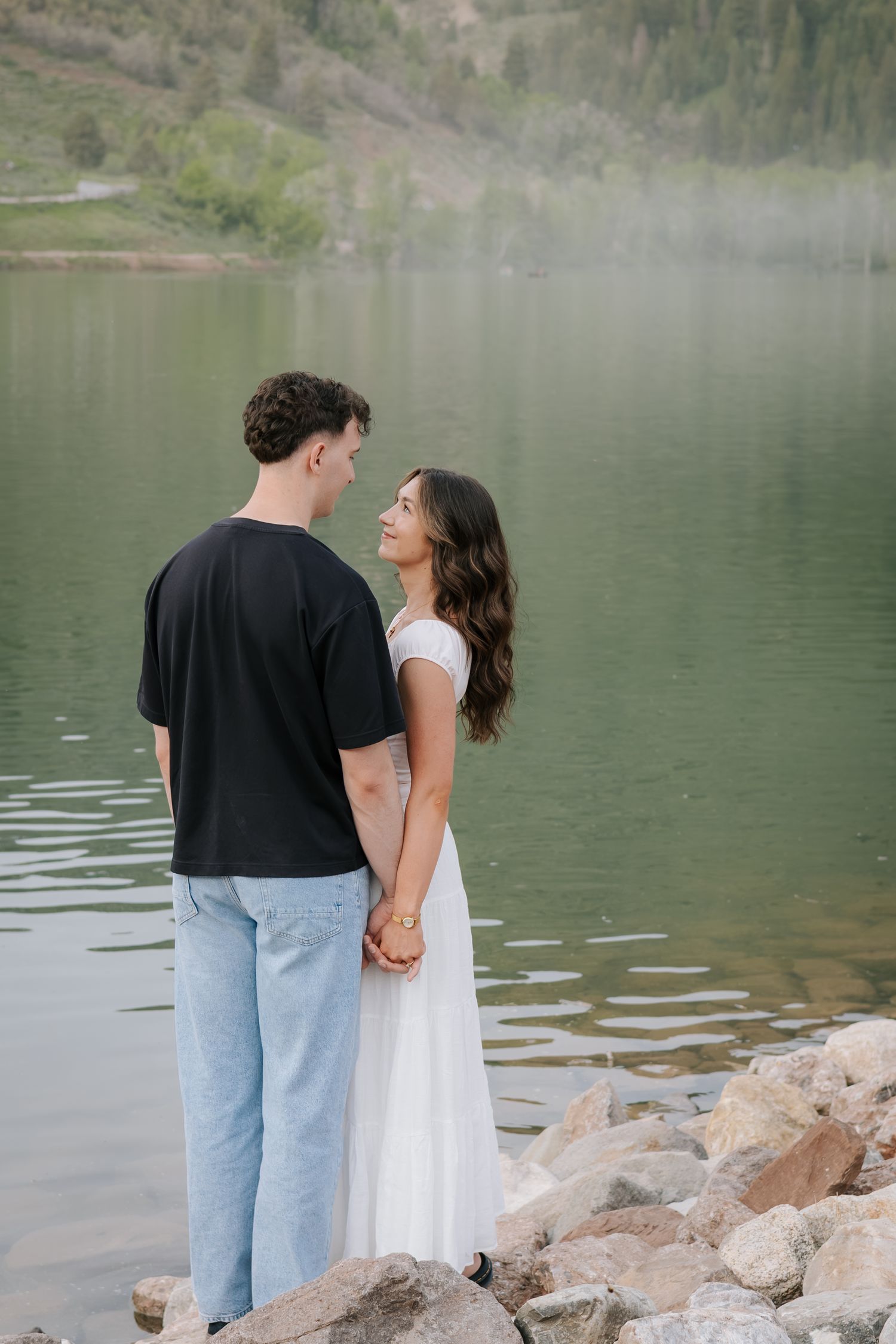 A couple stands together by a serene mountain lake surrounded by rocky shores.