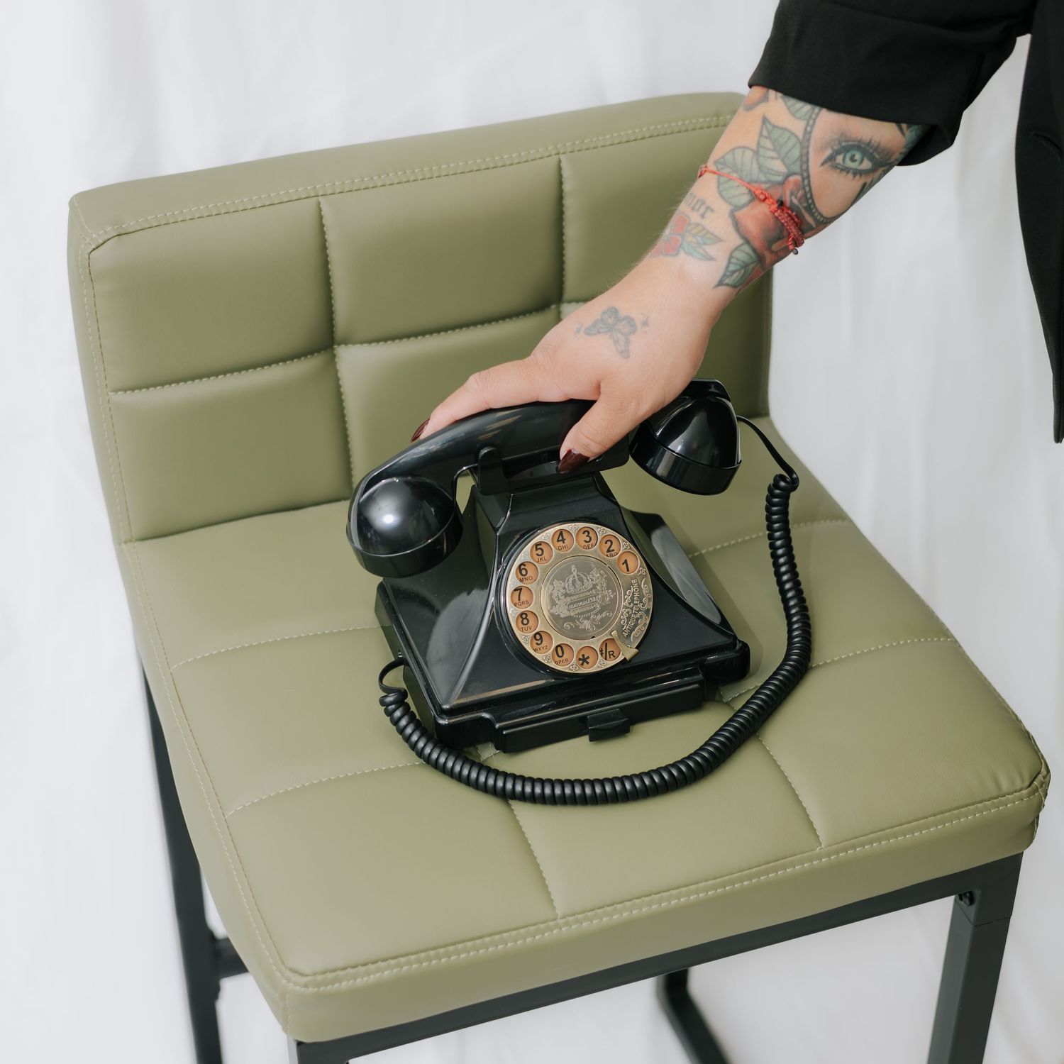 Tattooed hand placing vintage black rotary telephone on olive green leather chair.