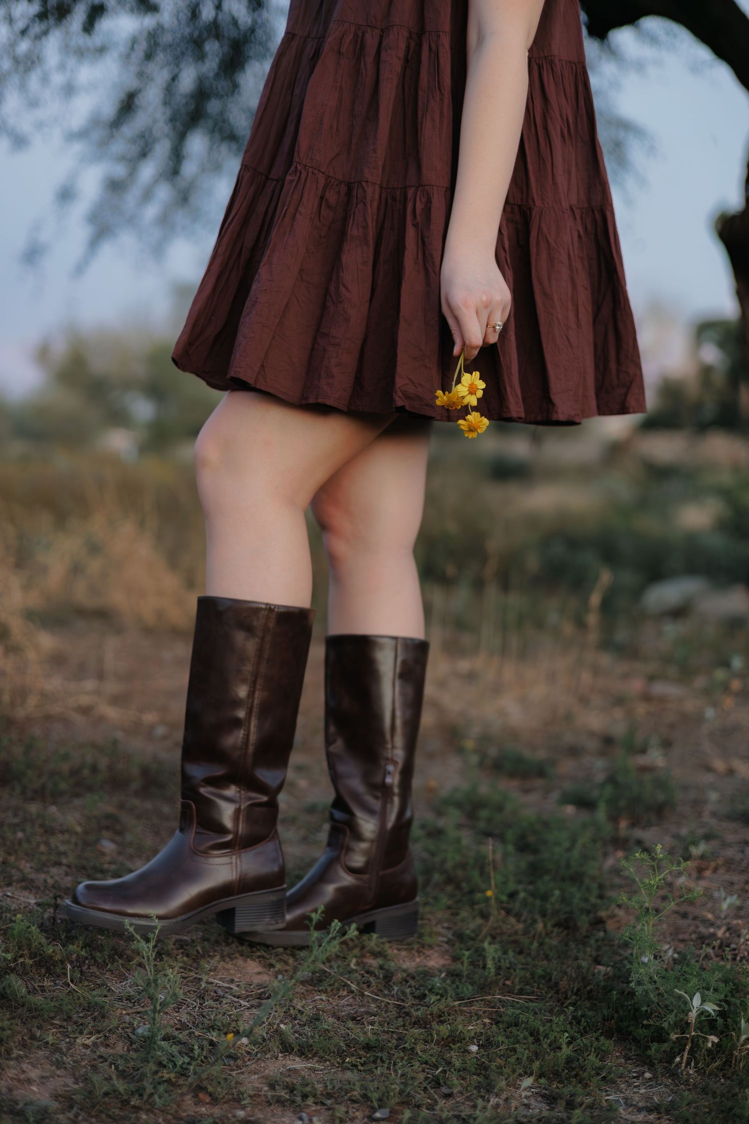 Brown leather boots and burgundy dress hem shown against desert ground with small yellow wildflowers.