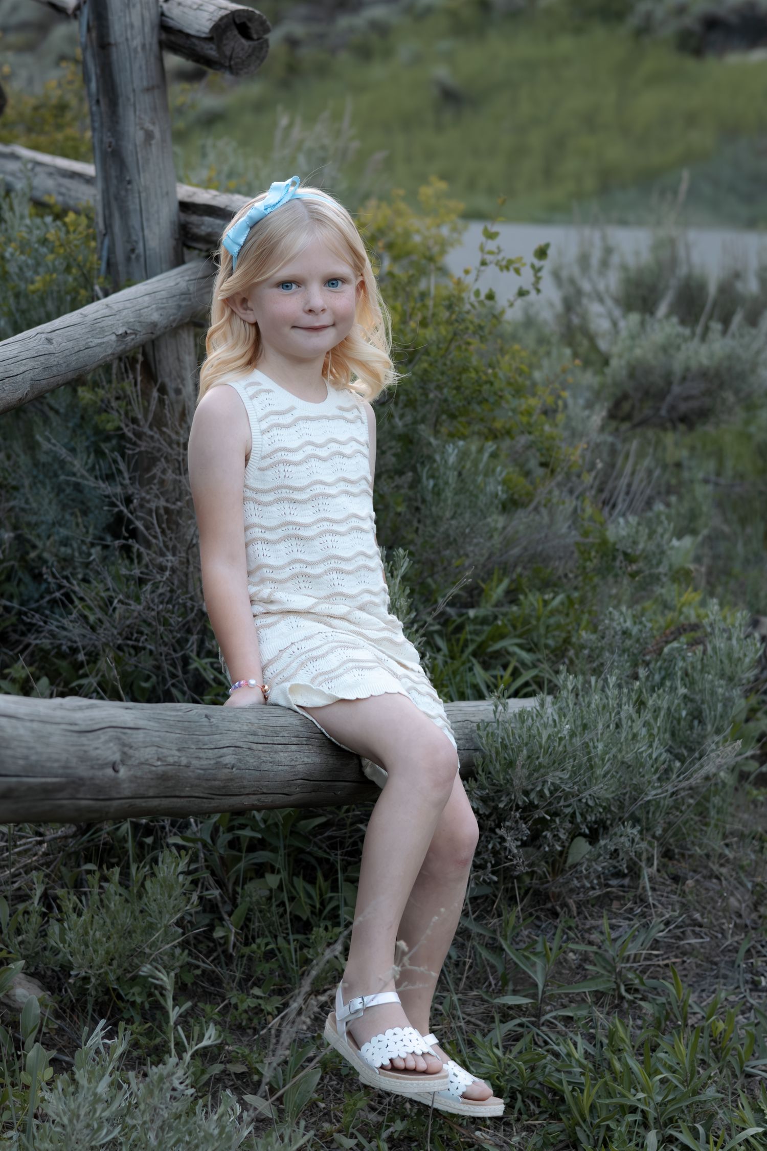 Young girl in white dress sitting on rustic wooden fence surrounded by greenery.