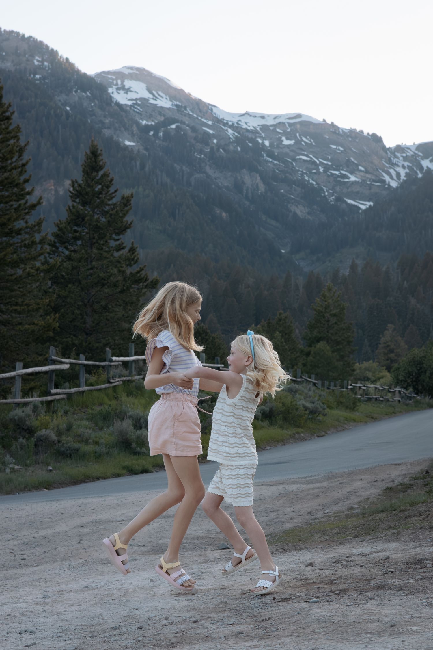 Two children playing and running on a mountain path with snow-capped peaks in the background.