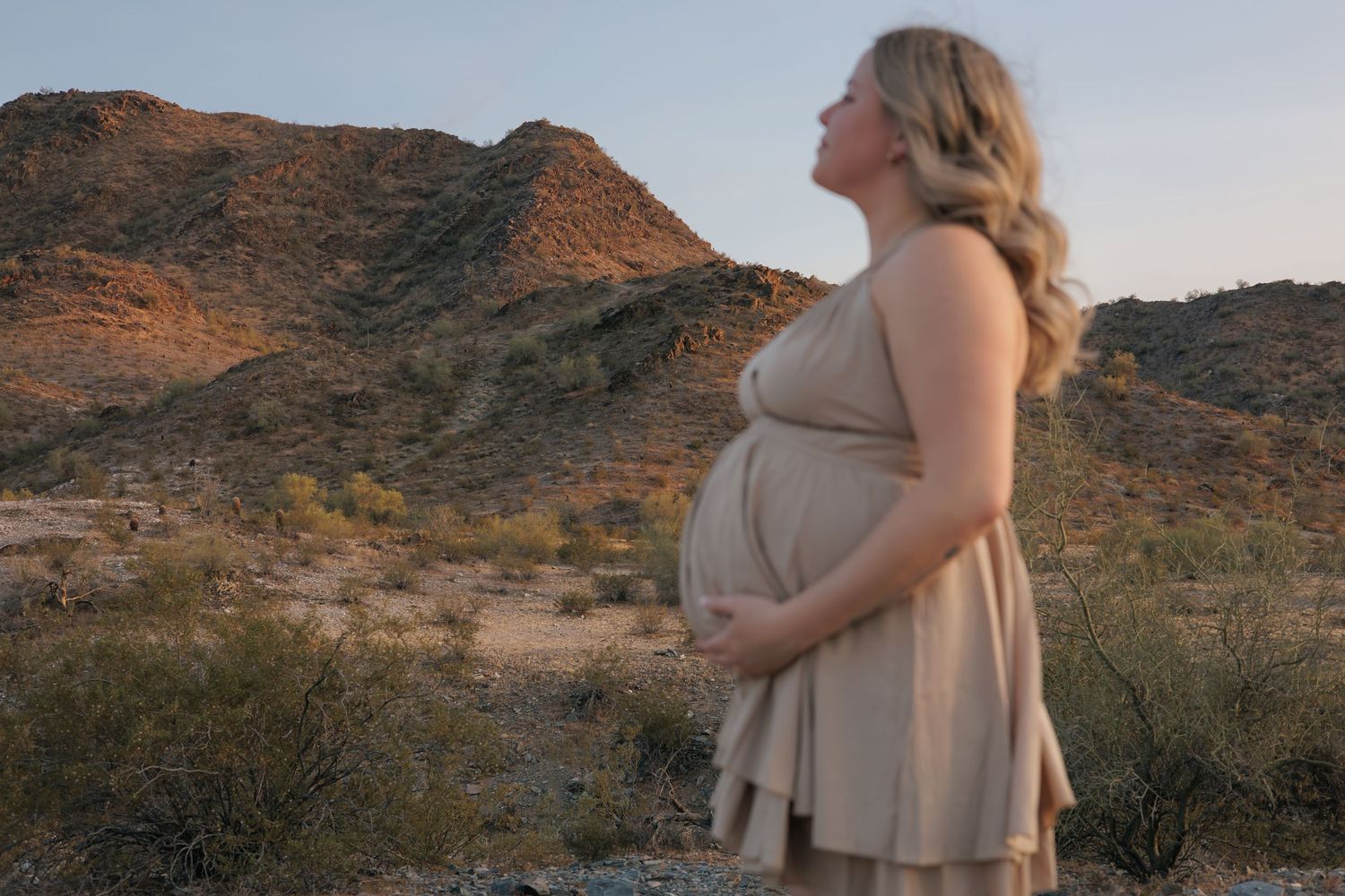 A pregnant woman in a beige dress stands in a desert landscape at sunset with rugged mountains in the background.