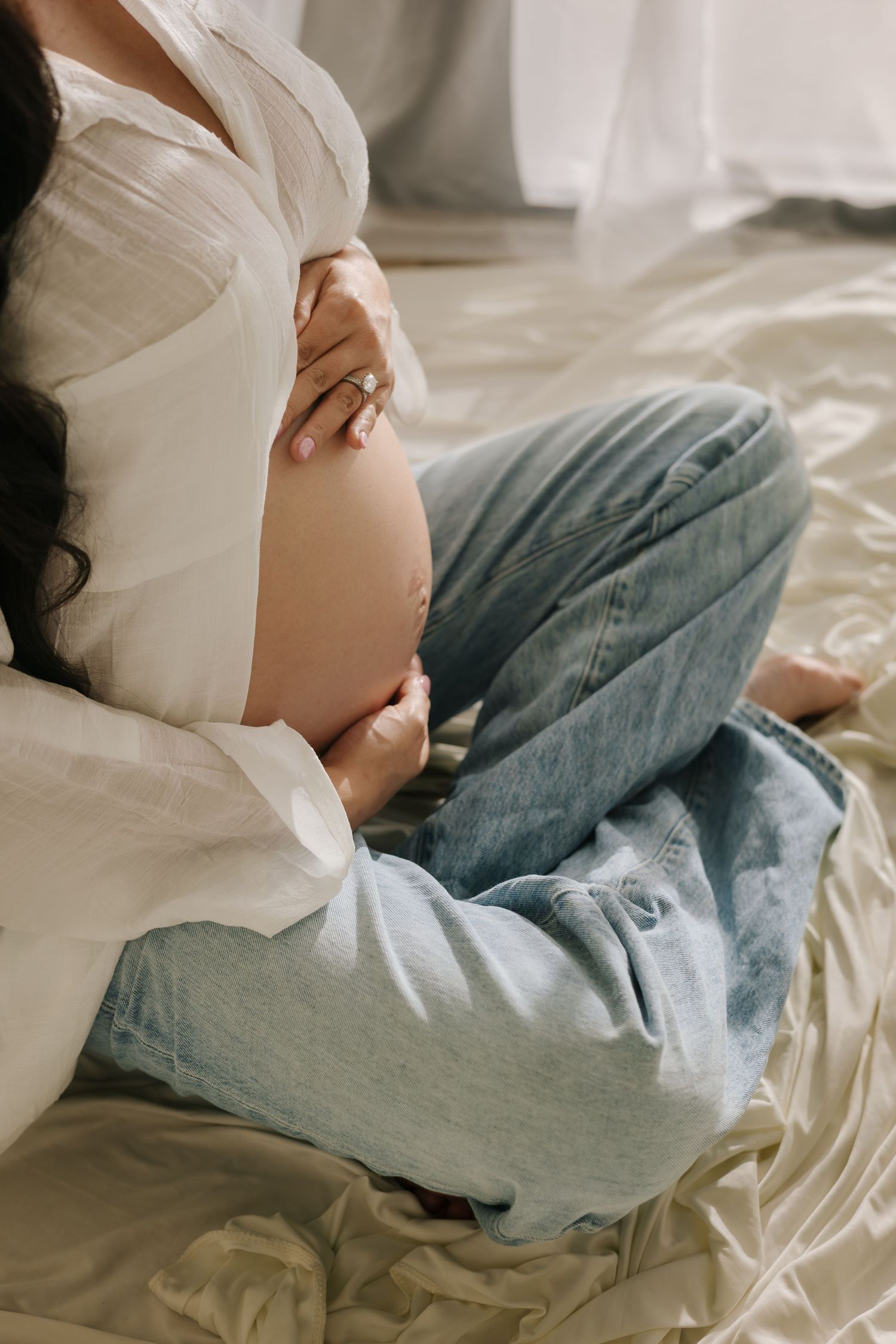 Intimate maternity photos showing pregnant belly in casual denim and white shirt on bed.