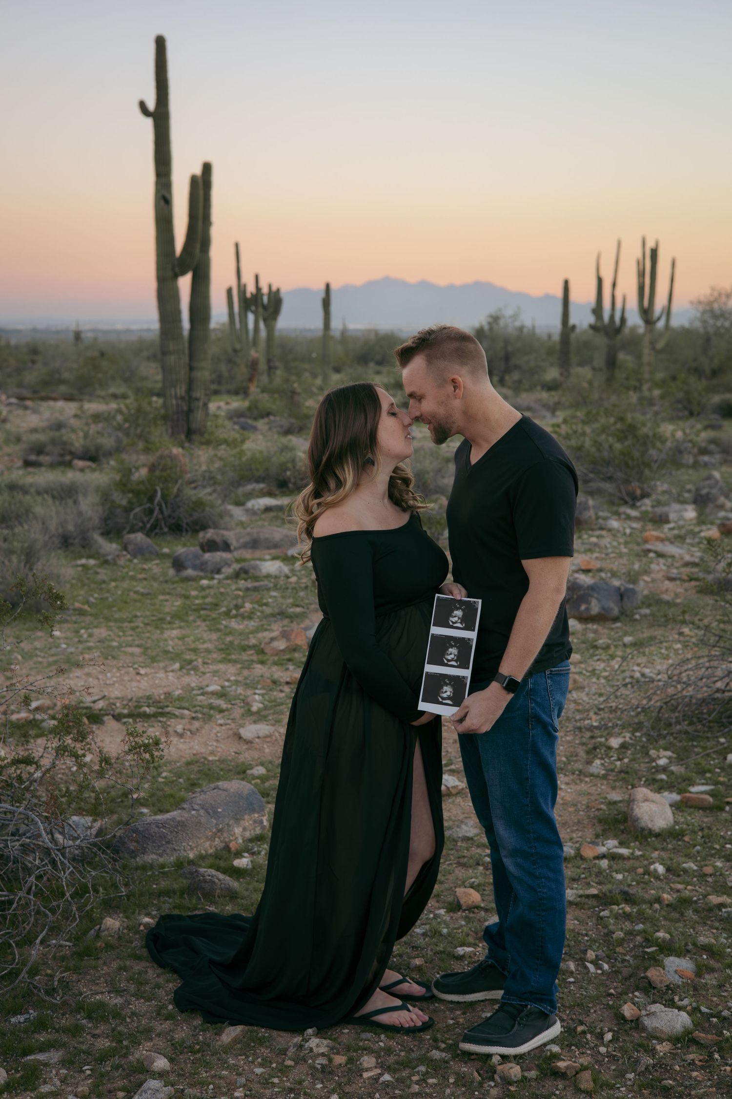 Couple sharing intimate moment while holding ultrasound strip in desert with saguaro cacti.