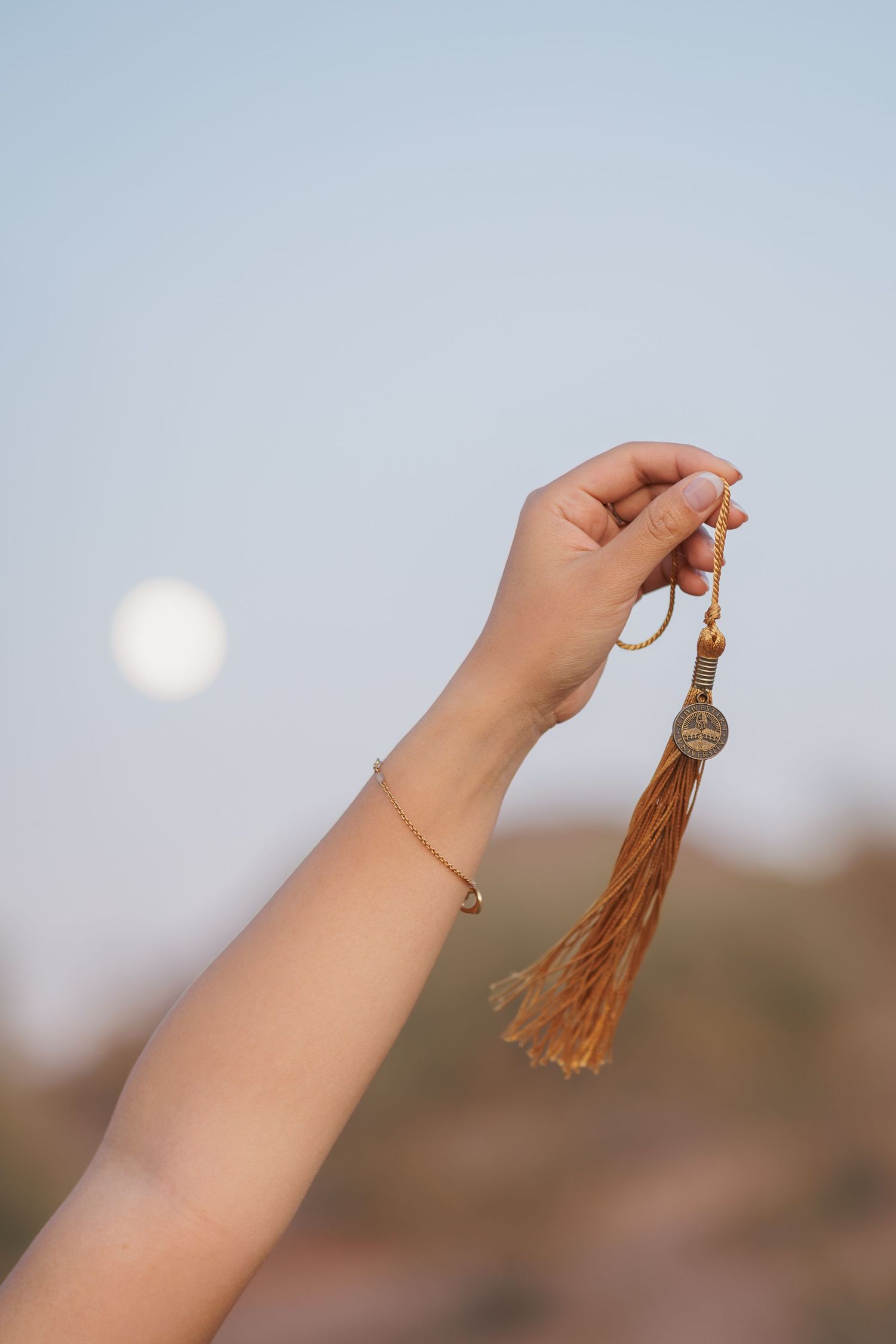 Hand holding a gold tassel charm bracelet against a blue sky with a full moon in the background.