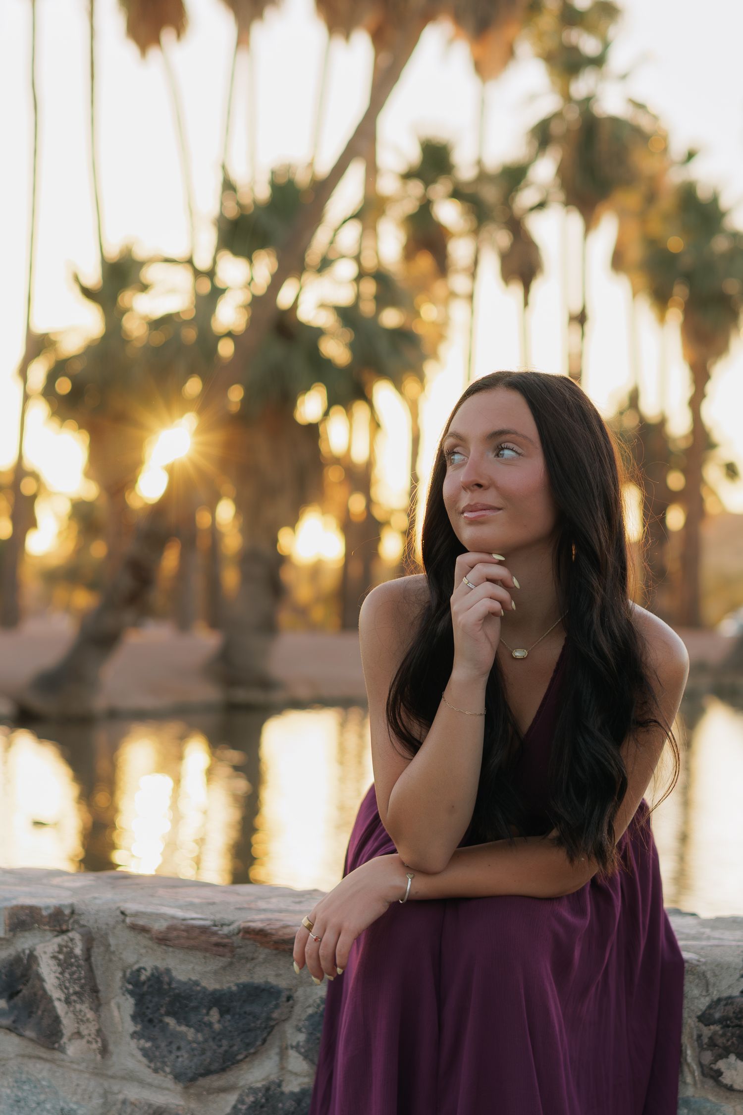 A young woman in a purple dress poses at sunset near a reflective pond with golden palm trees in the background.