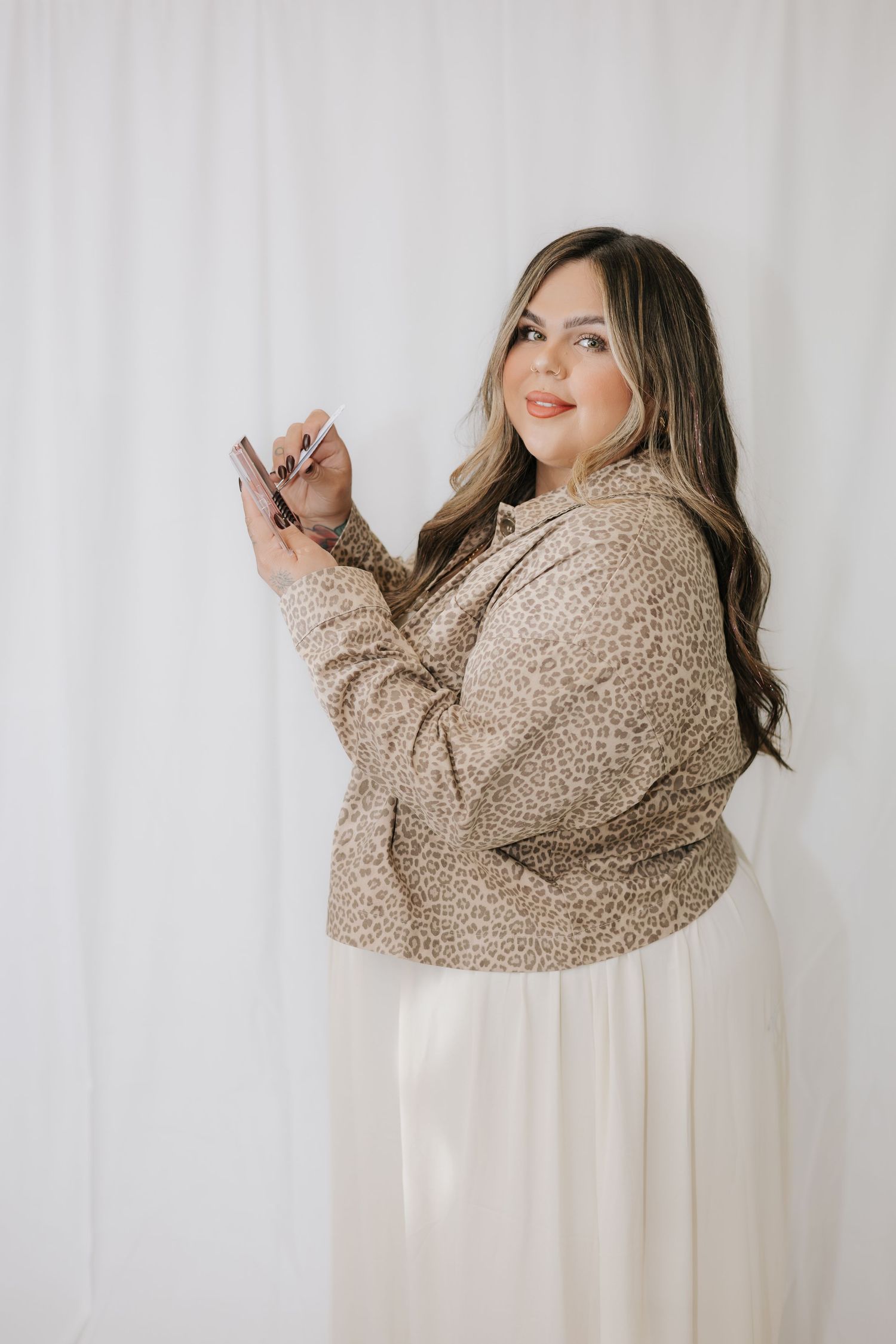 Woman in beige leopard print jacket posing playfully against white studio backdrop.