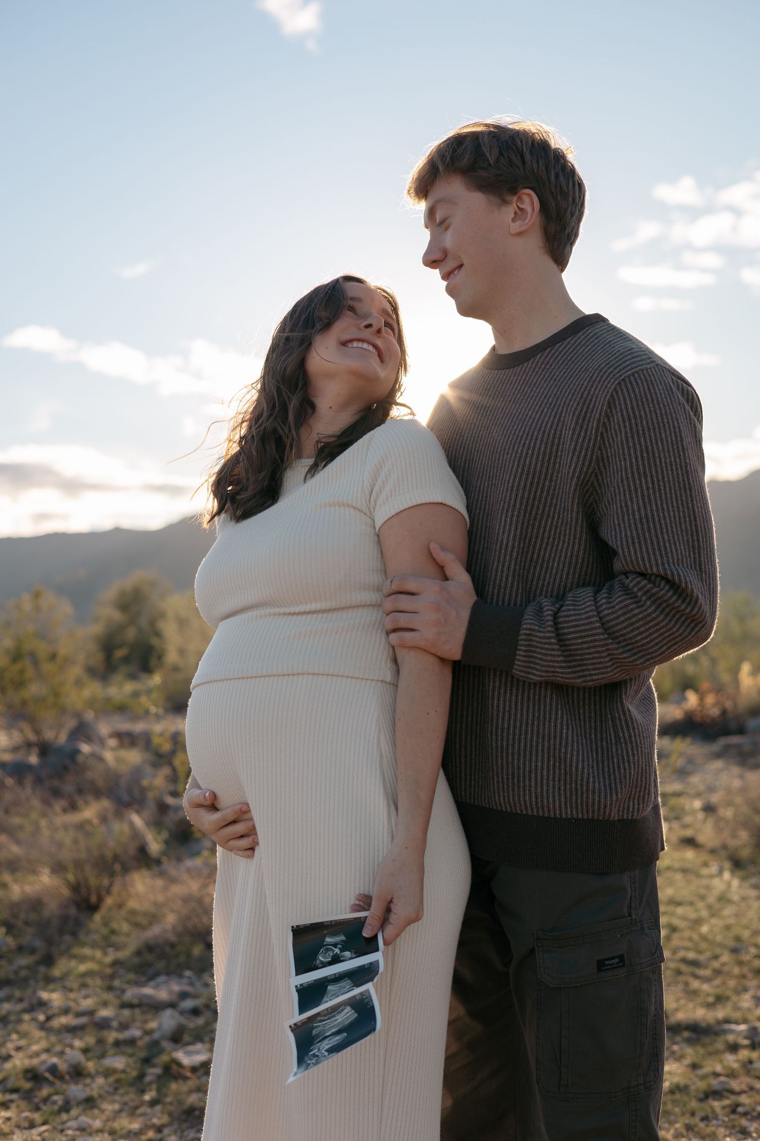 Couple sharing tender moment while holding ultrasound photos in outdoor setting.