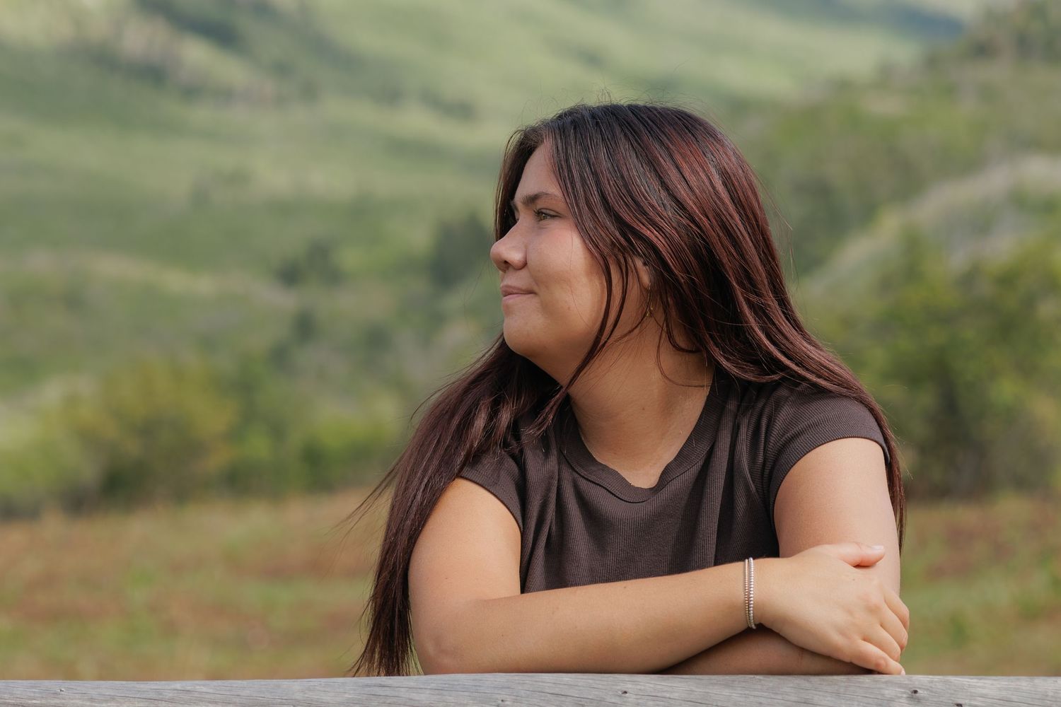 Young woman with long dark hair resting arms on a fence, gazing into lush green hillside landscape.